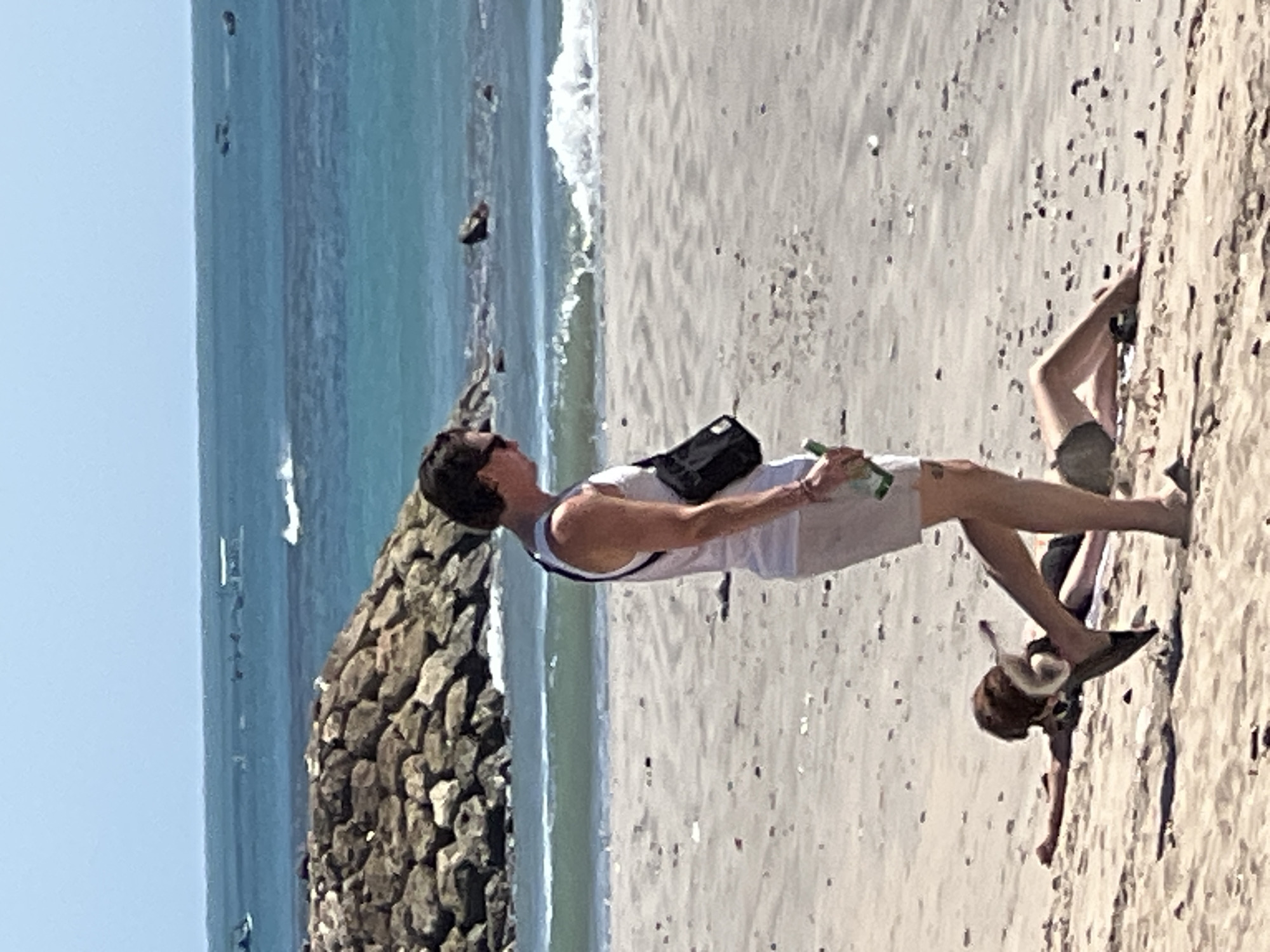 A medium shot captures a man walking on a sandy beach in Kuta, Indonesia. He is wearing a white tank top and light-colored shorts, with a black fanny pack slung across his chest. In his right hand, he holds a green bottle. In the foreground, on the left, a person is lying down on the sand with their head and torso covered by a brown object, possibly a dog. To the right of this person, another individual is lying on their back with their legs in the air, wearing dark shorts and a dark top. In the background, the turquoise ocean stretches out to the horizon under a clear blue sky. A rocky breakwater is visible along the coastline. The beach is scattered with small stones and debris.