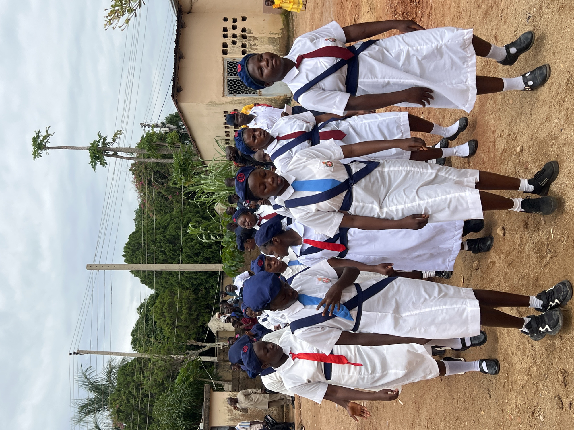 In the city of Jos, Nigeria, a group of young people, predominantly female, are parading outdoors in what appears to be a school or organized event. The subjects are dressed in white uniforms, many featuring blue berets and neckties in red, blue, or maroon. Some individuals have a blue sash across their chest, adorned with a crest. They are walking on a dirt path lined with buildings and lush green trees under a cloudy sky. The buildings have a tan exterior, one with a corrugated metal roof and barred windows. In the background, utility poles with wires suggest the presence of infrastructure. The overall scene captures a formal procession within an outdoor urban environment.