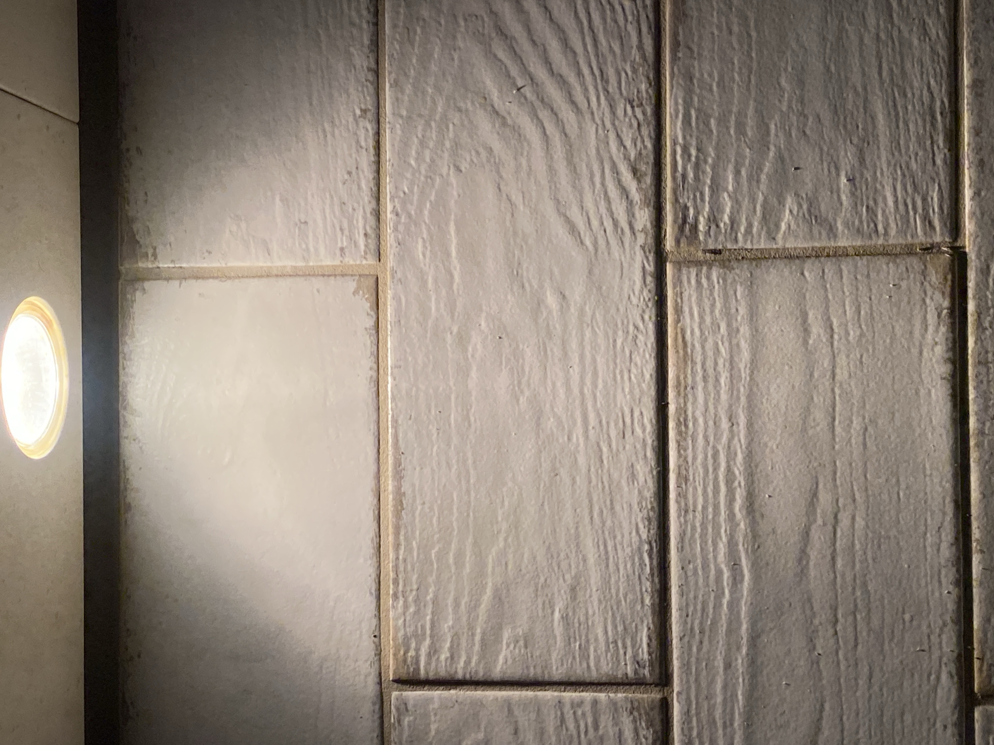 A close-up, eye-level, full shot shows a textured, light-colored tiled wall illuminated by a recessed ceiling light. The tiles are rectangular and arranged in a brick pattern. The grout lines between the tiles are a slightly darker shade of tan. The texture of the tiles appears to be somewhat rough and irregular, with visible striations that mimic wood grain. Some minor imperfections, such as small dark specks, are present on the tile surfaces. The lighting creates a bright, illuminated area directly beneath the light fixture, with the illumination fading as it moves down the wall, creating shadows and highlighting the texture of the tiles. The top edge of the image shows a dark band above the tiles, likely part of the cabinet or countertop. The scene is indoors, and there are no people or any other objects visible. The location in Chișinău, Moldova, cannot be determined from the provided image.