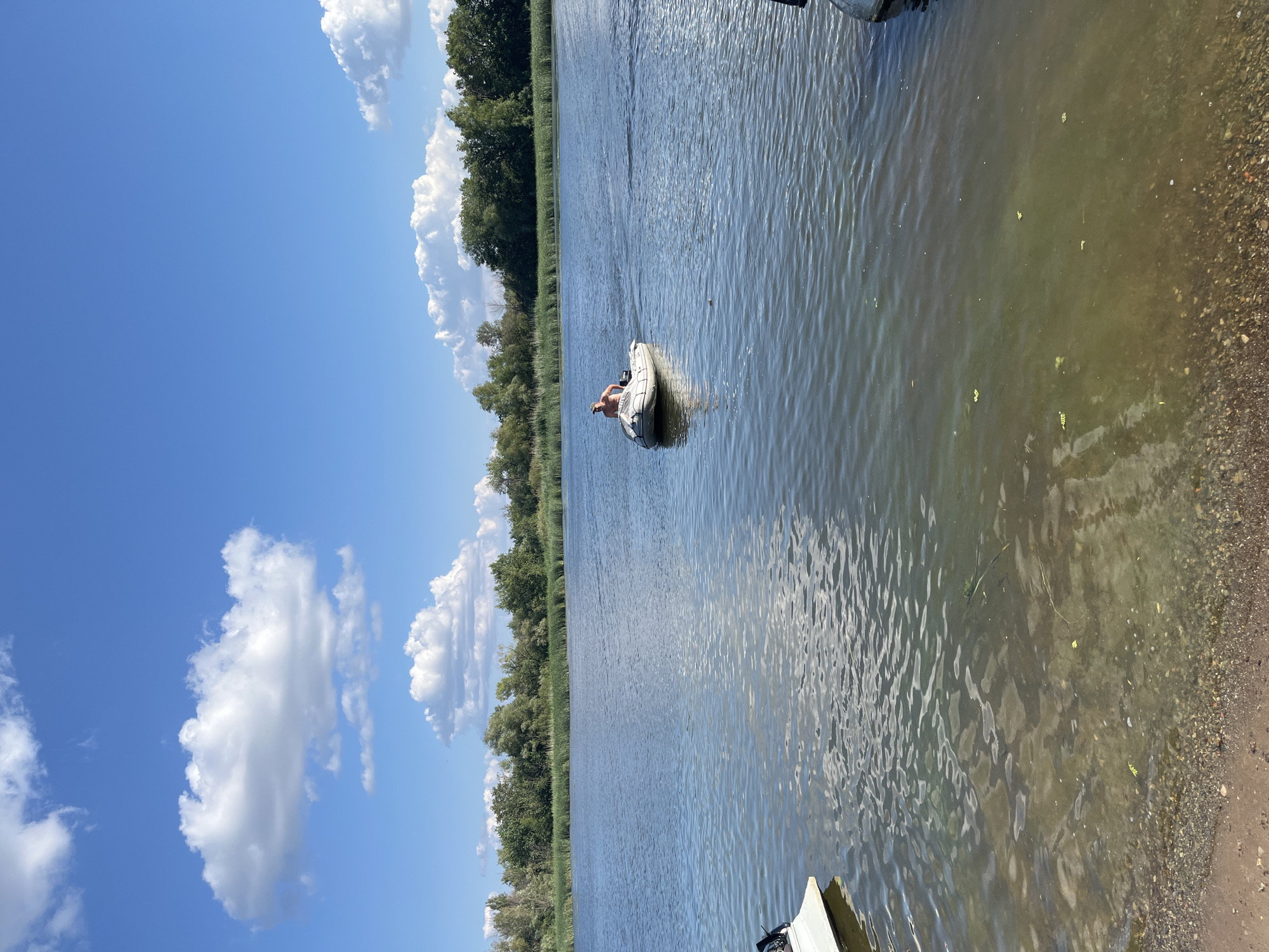 The image depicts a wide, calm river under a bright blue sky with fluffy white clouds. A person is seated in a small inflatable boat in the middle of the river. The boat is white and appears to be powered by an outboard motor, which is visible at the stern. The person is shirtless and wearing a baseball cap. The riverbanks are lined with dense green trees and reeds, suggesting a natural or semi-natural setting. The water in the foreground is shallow and clear, revealing a gravelly bottom and sandy shore. The overall scene conveys a sense of leisure and outdoor activity. There is no specific information in the image to identify the location as Saratov, Russia.