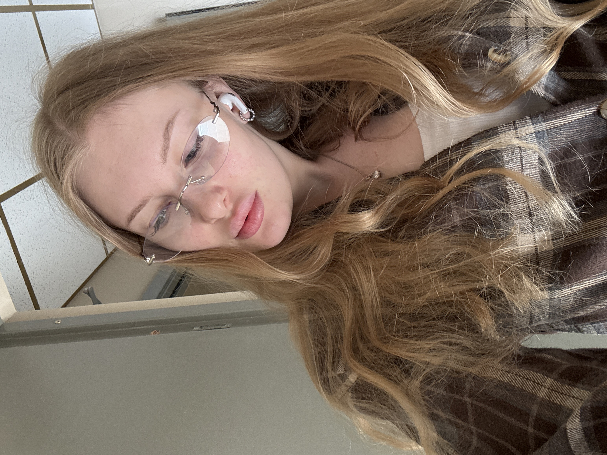 A young woman with long, blonde hair and wearing frameless glasses is the subject of this photograph. She is wearing an earbud in her left ear and a brown plaid shirt. She also has a silver necklace on. The background shows a portion of a gray surface and a tiled wall. The location of the photograph is not apparent. The city of Perm, Russia cannot be confirmed or denied.