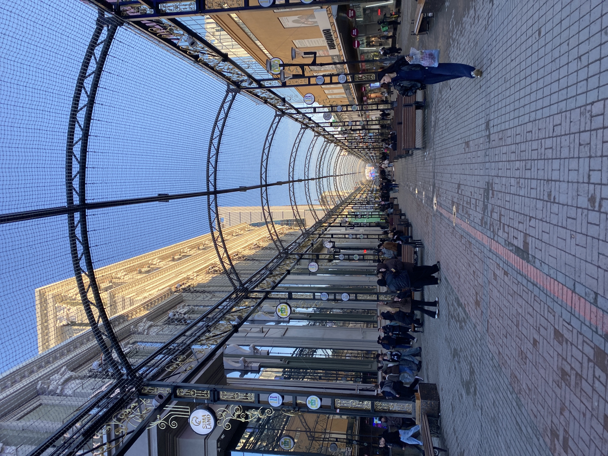 The image shows a long, narrow pedestrian street in Yekaterinburg, Russia. The street is lined with buildings on both sides, covered by an arched metal structure covered with netting. Benches line the street. People are sitting and walking along the street. The street surface is paved with brick. Storefronts and signage are visible on the buildings.