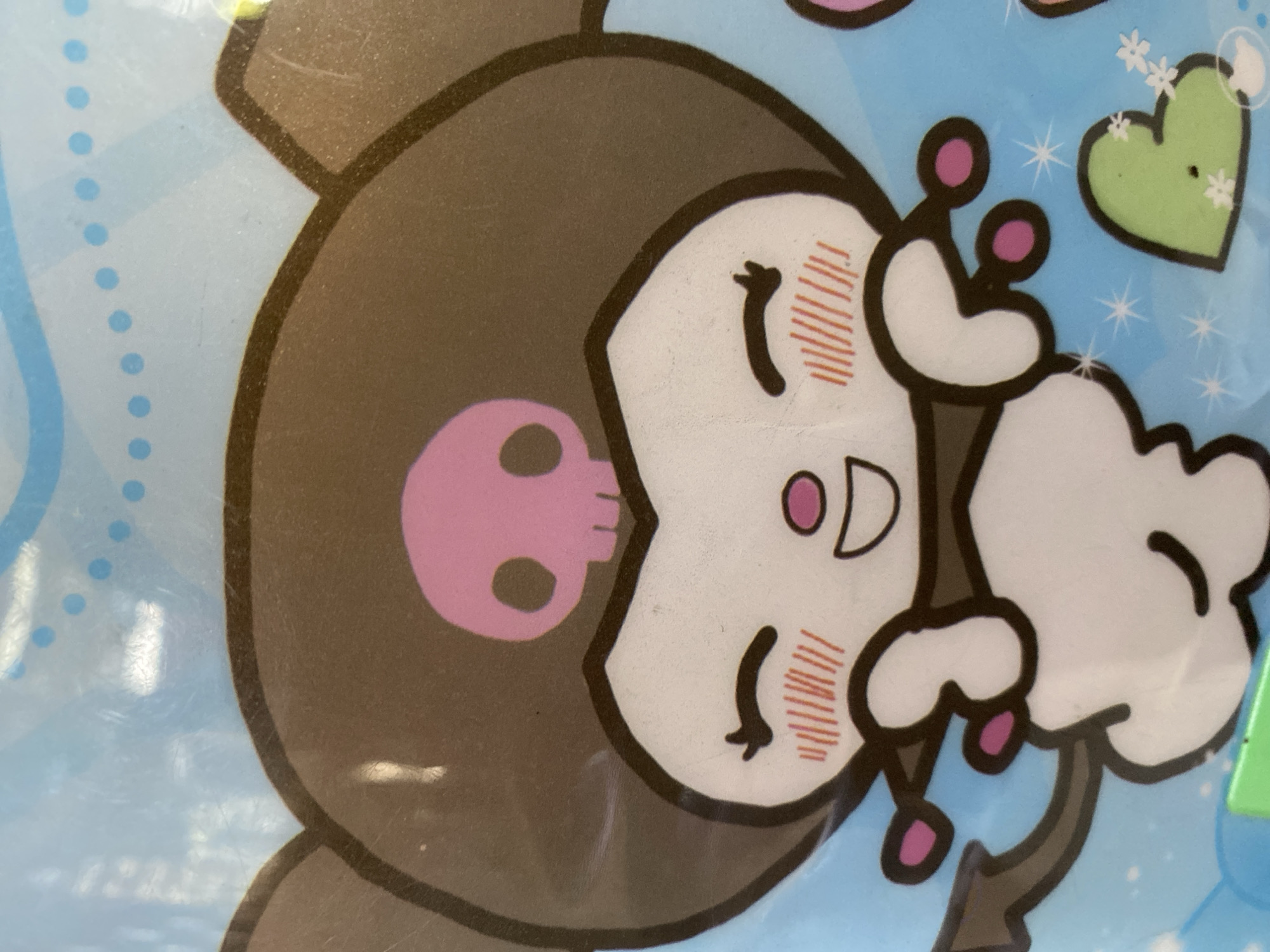 The image is a close-up of a cartoon character, possibly from a Sanrio franchise, against a light blue background with small blue polka dots and white starbursts. The character has large brown ears, a brown hood with a pink skull emblem on the forehead, and a white face with closed, smiling eyes and rosy cheeks. The character is depicted with its arms raised, holding small pink spheres at the end of each hand, which are attached by thin brown lines. A dark brown, arrow-shaped tail with a pink tip extends from the character's lower body, which is mostly white. To the lower right of the character is a light green heart shape adorned with small white flowers. The image appears to be a photograph of a printed surface, exhibiting some glare and minor surface imperfections. There are no identifiable people or specific locations in Ta', Indonesia.