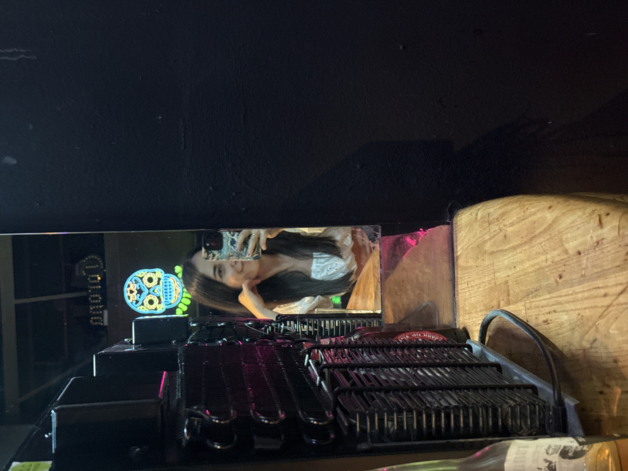 The image shows a woman taking a selfie in a mirror. She is wearing a white top with lace detailing and has long dark hair. She is leaning on her hand and smiling at the camera. In the mirror's reflection, a neon skull decoration is visible behind her. The background is dark, with what appears to be a black wall on the right side of the frame. A black, ribbed object, possibly a piece of electronic equipment or a cooling unit, is to the left of the mirror. A wooden surface, likely a bar or tabletop, is in the foreground. The scene appears to be set in a bar or entertainment venue. There is no indication of the location being Barcelona, Spain.