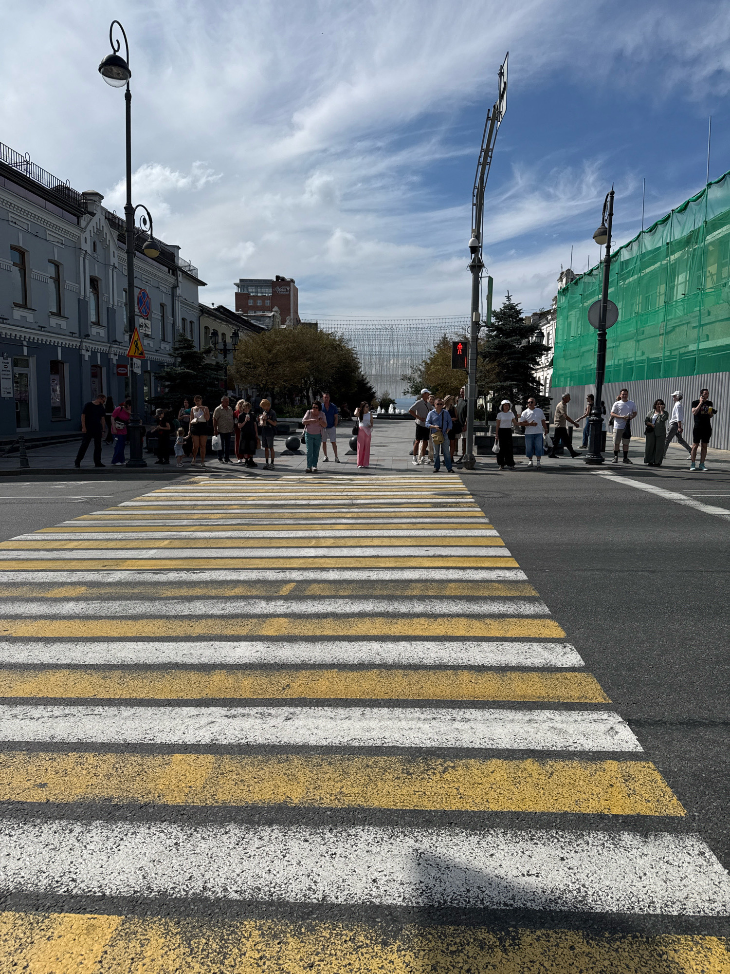 The image shows a crosswalk in Vladivostok, Russia. A group of people is waiting to cross the street. The buildings on the left are old, with traditional architecture. The building on the right is covered in green netting, suggesting it is under construction or renovation. The sky is cloudy, with patches of blue visible. The street is paved with asphalt. The crosswalk is marked with white and yellow stripes. A traffic light is visible, showing a red pedestrian signal.