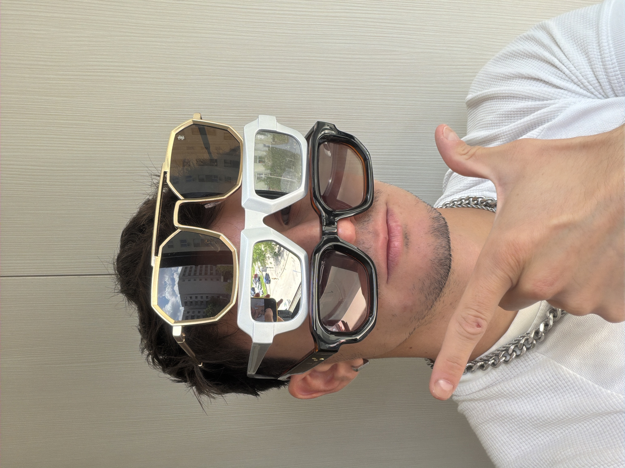 A person is wearing three pairs of sunglasses on their face. The sunglasses have different frame styles: gold, white, and black. The lenses reflect the surroundings, showing buildings and blue sky. The person is also wearing a white waffle-knit shirt and a silver chain. Their hand is gesturing downward with their index finger. The background is a light-colored wall. Without additional information, it is not possible to determine if the location is in Sofia, Bulgaria.