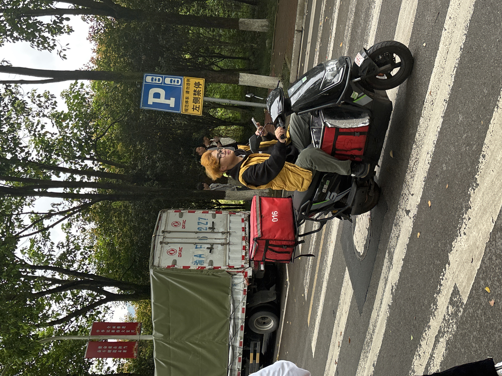 A man with orange hair is sitting on a black scooter on a crosswalk in Jiangjin, China. He is wearing glasses, a black jacket, and a yellow vest. He is holding a phone in his left hand. The scooter has a red delivery bag attached to the back with the number "016" on it. Behind the scooter is a white truck with a green tarp covering its cargo bed. The truck's license plate is "渝AF62Z7". There is a parking sign and a sign that reads "规范停车 请勿违例 左侧禁停" in Chinese. There is also a red sign with Chinese text that says "中华民族伟大复兴 全面推遣" and "奋斗创造未来 使命重在担当" (Rejuvenation of the Chinese nation, comprehensively promoted; Strive to create the future, The mission lies in the responsibility). The background includes trees and other people. The brand name "绿源" is displayed on the scooter.