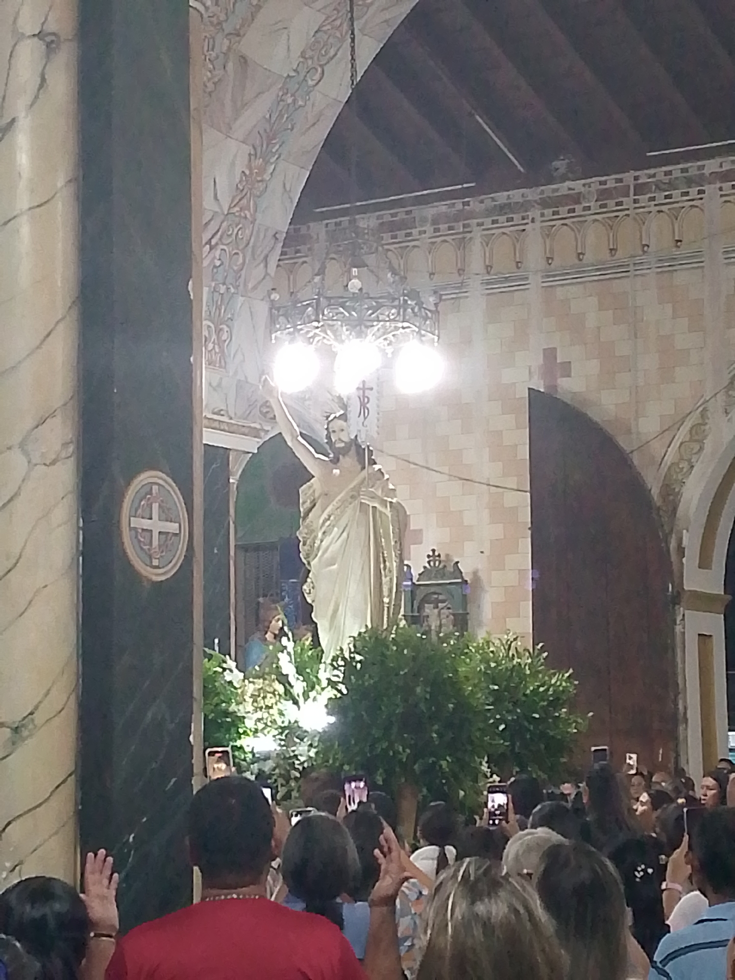 The image depicts the interior of a church in Tinaquillo, Venezuela, during what appears to be a religious event. A large statue of Jesus Christ is the focal point, situated in front of a large light fixture and surrounded by greenery. A crowd of people stand before the statue, many holding up their smartphones, seemingly taking photos or videos. A statue of a saint can be seen at the base of the statue of Jesus Christ. The church's architecture is visible, featuring arched doorways, detailed archways, and brick walls. A man in a red shirt is standing in the crowd with his hand raised, seemingly greeting the statue.