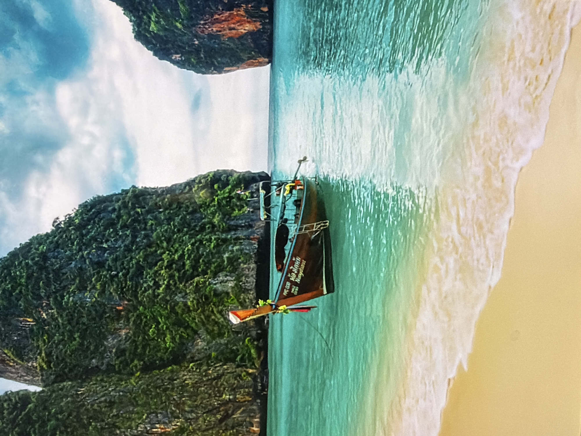 The image displays a vibrant tropical coastal scene, vertically oriented and showing a longtail boat in turquoise waters next to a sandy beach and towering limestone karsts.
**Subjects:** The primary subject is a traditional Thai longtail boat, dark in color, with a prominent engine structure at the stern and decorative floral garlands on its prow. The boat is positioned in clear, shallow, light blue-green water. To its right (top in the original image), a pristine sandy beach with gentle white waves breaking on the shore is visible. On the left and top (bottom and left in the original orientation) are large, steep limestone islands or karsts, densely covered in lush green vegetation. The sky above is bright blue with scattered white clouds.
**Activity or event taking place:** The boat appears to be moored or gently approaching the shore, likely for passenger drop-off or pick-up, typical of tourist activities in such a location. The calm water suggests a peaceful moment.
**Notable details:**
* **Time of day:** It is bright daylight, indicative of late morning or early afternoon, with ample sunlight illuminating the scene.
* **Weather:** The weather is clear and sunny, with a few soft clouds in the sky.
* **Location cues:** The distinctive longtail boat and the dramatic limestone karsts are strong indicators of a Southeast Asian location, most likely the Andaman Sea region of Thailand (e.g., Phi Phi Islands, Krabi).
* **Emotions:** The scene evokes feelings of serenity, natural beauty, and a popular tourist destination.
**Visible text:** On the side of the boat, the following text is visible:
* "P.P.C. 1026"
* "หอยทาก" (Thai script, translating to "Snail" or "Oyster")
* "Nonghant" (likely a boat name or owner's name in English transliteration)