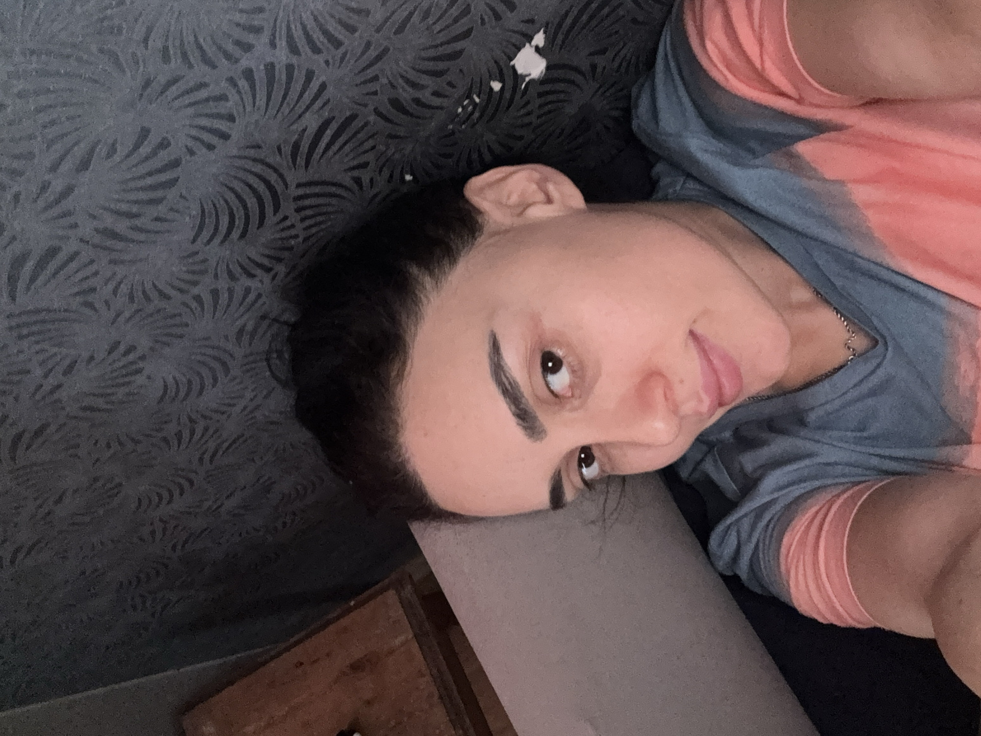 A selfie taken from a low angle depicts a person lying on a patterned, dark blue-grey surface. The person has dark hair and fair skin. They wear a multi-colored shirt with blue-grey on top and pink on the sides. The person's head rests on a grey pillow or cushion. The environment includes a textured wall and a glimpse of what appears to be a wooden cabinet or furniture along the bottom edge of the frame.
It is not possible to identify the location of this photograph as being in the city of Tbilisi, Georgia based on the context provided.