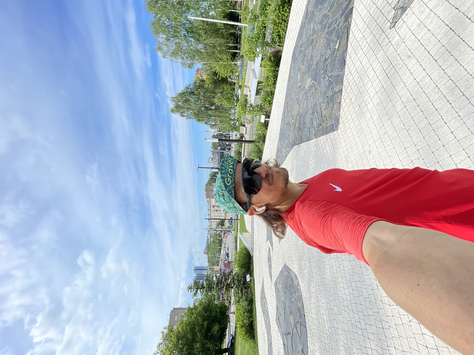 A medium shot, taken from a slightly low angle, captures a person taking a selfie outdoors on a sunny day. The subject is a male with light brown hair and a beard, wearing a bright red short-sleeved Nike shirt with the white swoosh logo visible on the left chest. He has sunglasses on and a green patterned cap, possibly a cycling cap, with the word "Gravel" clearly visible, along with partial words like "Graveling," "adventure," and "bike." There's also some text that appears to be "СОКРАЯ" on the cap. He has white wireless earbuds in his ears and a noticeable tan line on his arm where his sleeve ends.
The person is looking slightly upwards and to his right, away from the camera, suggesting he is enjoying the weather or surveying the surroundings. The scene unfolds along a paved pathway, primarily light-colored square tiles, with sections of darker, irregular stone mosaic patterns.
In the background, a vibrant green urban park or promenade is visible, filled with numerous deciduous trees (some resembling birches) and bushes. Further in the distance, a road with several parked cars and distant city buildings can be seen under a bright blue sky dotted with scattered, wispy white clouds. The overall impression is a pleasant, clear daytime setting in an urban green space.