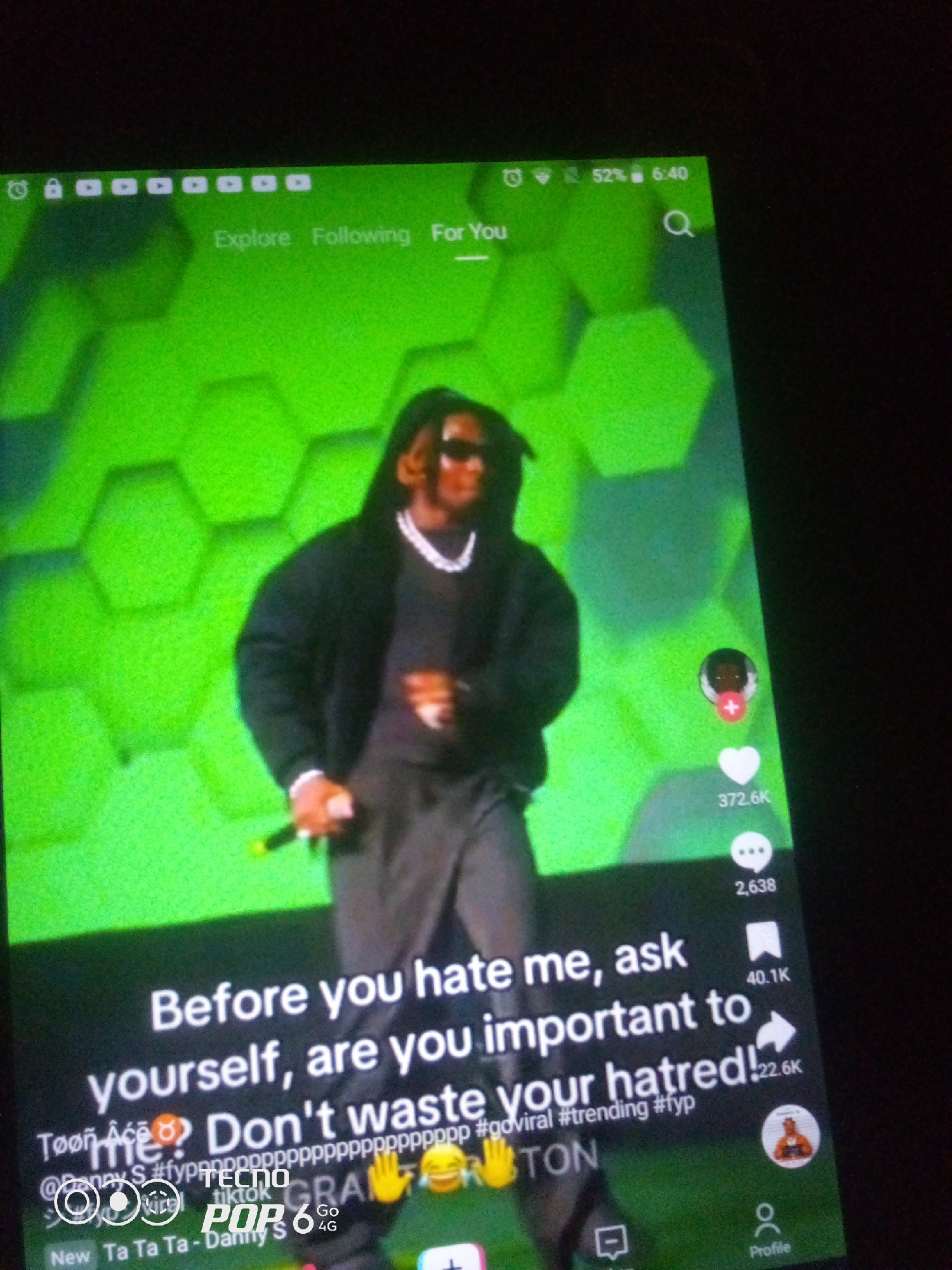A person wearing sunglasses and a black sweater is shown on a stage with a neon green background. They are holding a microphone and appear to be performing. The image is a screenshot of a social media video, likely TikTok, given the interface elements such as likes, comments, shares, and profile icons. The text overlay reads: "Before you hate me, ask yourself, are you important to me? Don't waste your hatred! #goviral #trending #fyp". Below this, the words "TECNOS POP 6 4G" are visible, indicating the phone model used to capture or display the video. The background features a repeating hexagonal pattern. The time displayed on the screen is 6:40 and the battery shows 52%. There are navigation icons at the top of the screen that read "Explore", "Following", and "For You". The location of Howayi, Nigeria, is not discernible from the image itself.