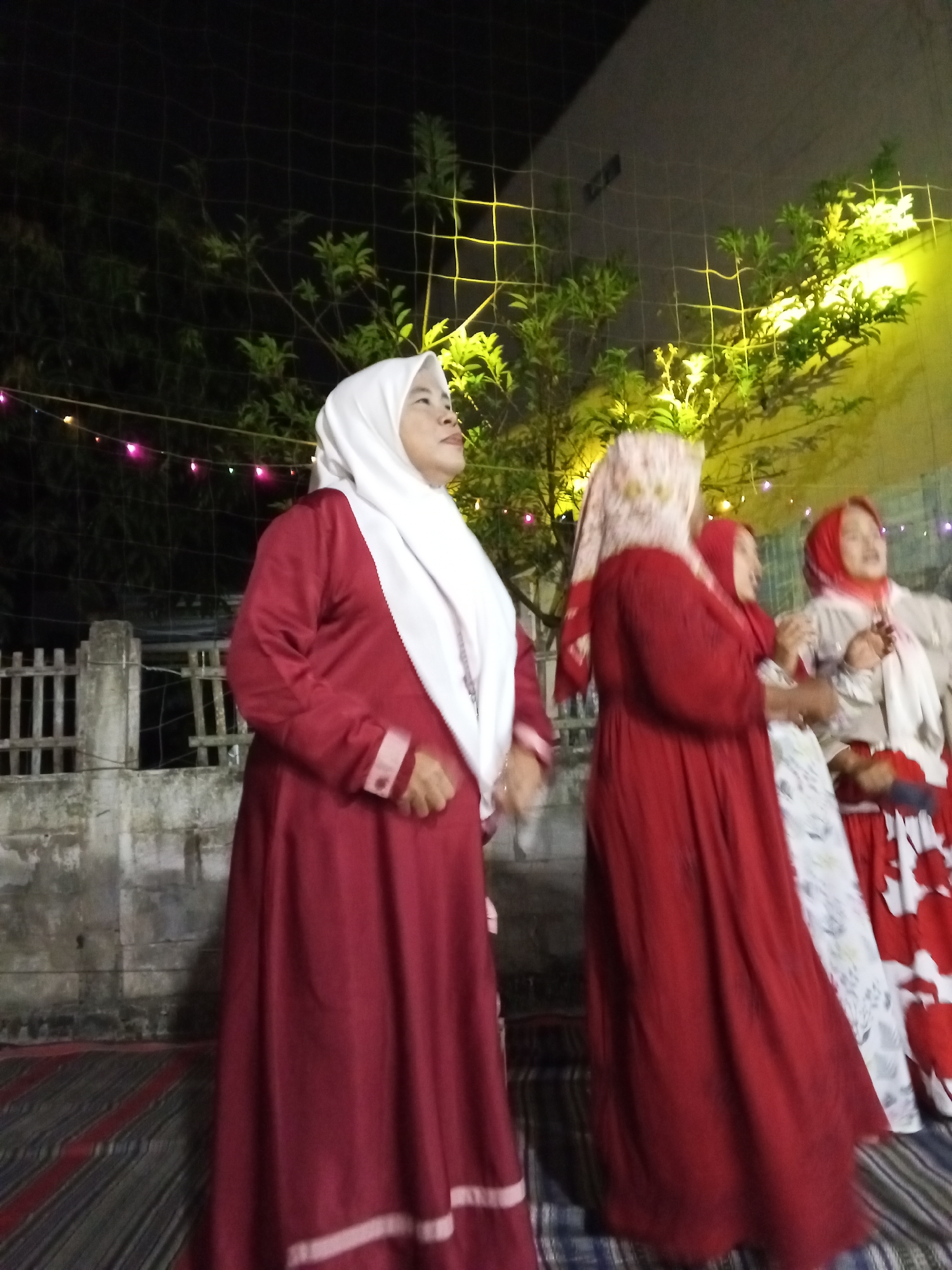 In the city of Ciruas, Indonesia, four women are seen outdoors at night, possibly during a celebration or gathering. The woman on the far left wears a maroon dress and a white hijab, looking upwards. To her right, another woman in a red dress and a patterned hijab faces away from the camera. A third woman in a red hijab and white dress is positioned between them, her head tilted. The woman on the far right, wearing a red headscarf, is partially visible, and she appears to be holding something. The background features a wall with a fence, trees, and decorative lights. A patterned rug is laid out on the ground. The lighting suggests it is nighttime, with some artificial illumination highlighting the scene.