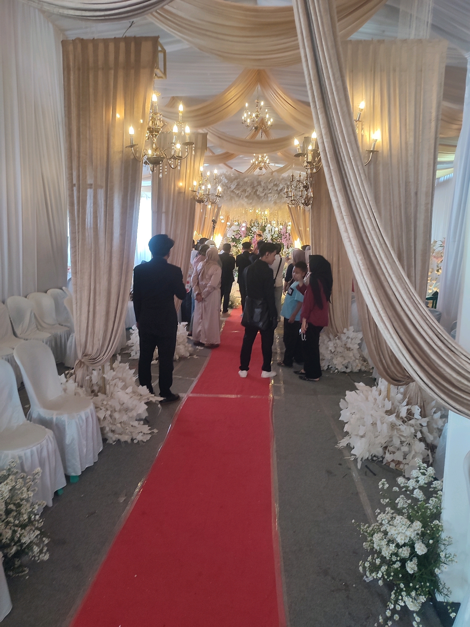 This image captures a wedding ceremony setup, likely an indoor event. A red carpet stretches down the center, leading towards a decorated altar or focal point obscured by a crowd of people. White chairs draped in covers are lined up on either side of the carpet, suggesting seating for guests. The ceiling is adorned with draped fabric in beige and white, creating a canopy effect, with ornate chandeliers hanging down, illuminating the space with warm light. Several people, dressed in formal attire, are present, some standing and conversing while others are positioned along the carpet. The overall ambiance is festive and celebratory, indicative of a wedding event. The location is identified as Kecamatan Haurgeulis, Indonesia.