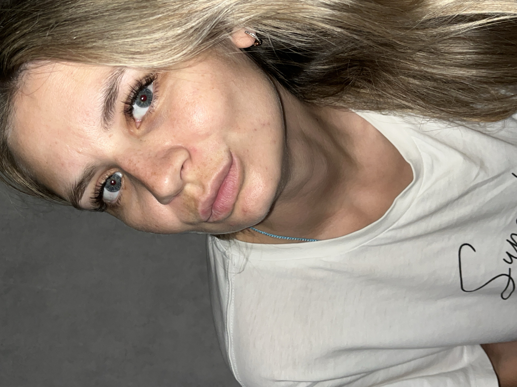 A fair-skinned individual with long, light brown hair and blue eyes looks directly at the camera, tilting their head to the right. Freckles are visible on their nose and cheeks. Their lips are pursed in a pout. They wear a white t-shirt, and a light blue beaded necklace is visible around their neck. A small earring adorns their right ear. The background is a plain, textured dark gray wall, indicating an indoor setting.