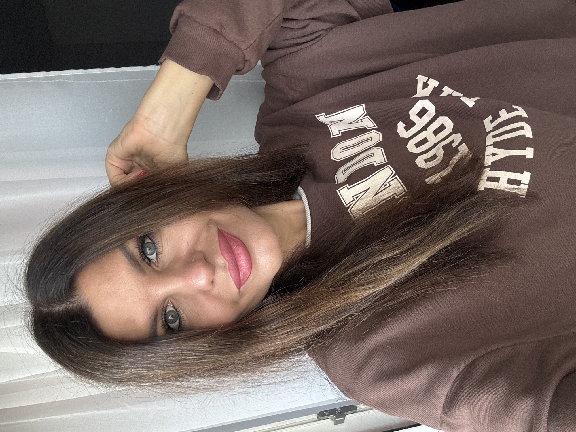 A woman is pictured in a selfie, looking into the camera with a slight smile. She has long, dark brown hair with lighter highlights, which she is casually touching with her right hand near the back of her head. She is wearing a brown sweatshirt with white lettering that appears to say "LONDON 1986 HYDE". Her makeup includes eyeshadow and pink lipstick. The background is blurred, showing a sheer white curtain on the left and a dark area on the right. The image is taken indoors, possibly in a residential setting. There is no indication of the city of Chaikovsky, Russia in this image.
