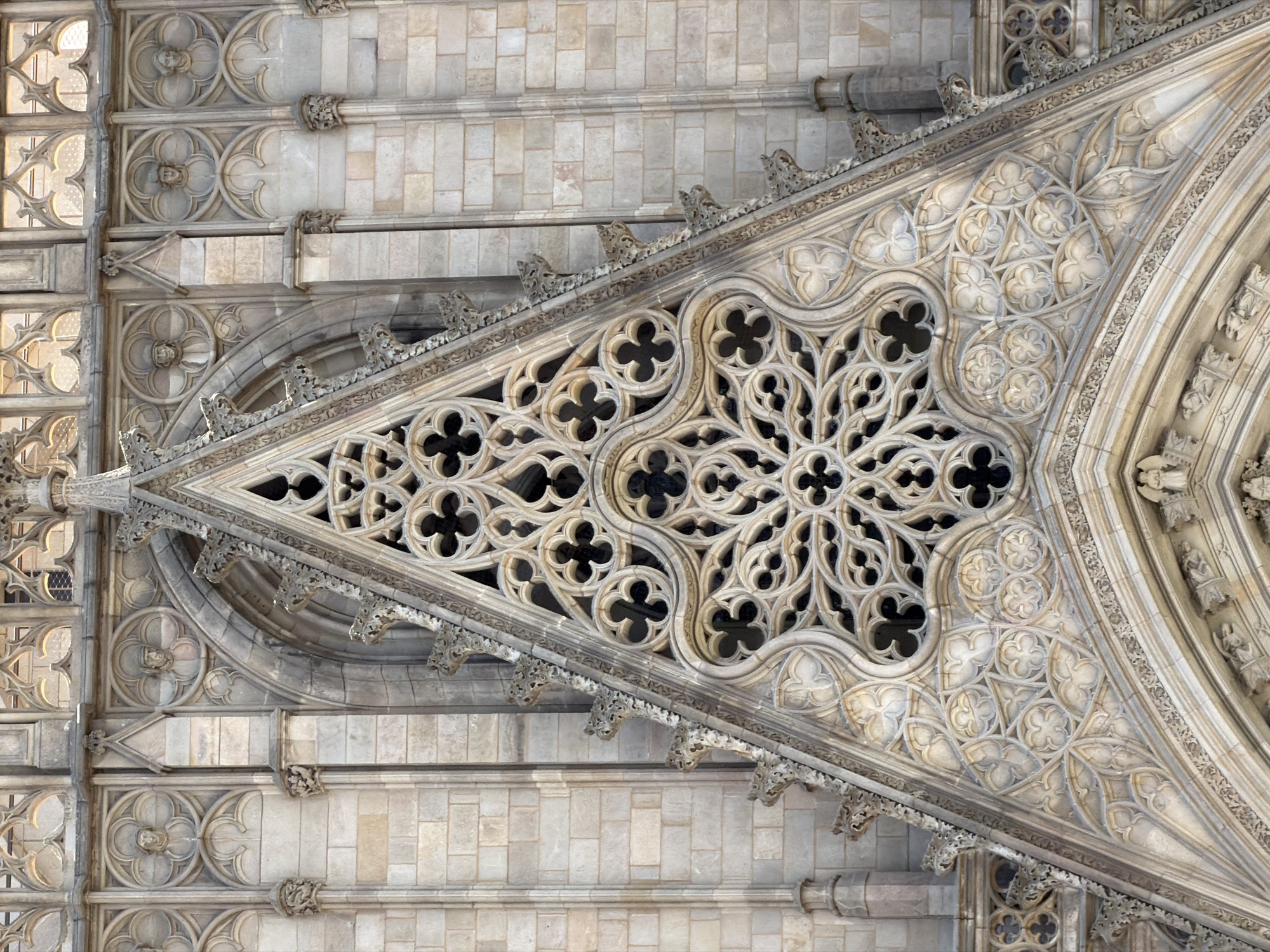 The image shows a close-up, eye-level, exterior shot of the facade of the Barcelona Cathedral in Barcelona, Spain. The facade is made of light-colored stone and features intricate Gothic architectural details. The central focus is a large, triangular gable adorned with elaborate stone tracery. This tracery includes a large rose window pattern composed of quatrefoils and other curvilinear shapes, set against a darker background. Below the rose window, the gable is decorated with a band of geometric patterns and trefoils. The surrounding stone walls are constructed of rectangular blocks and feature a series of smaller decorative elements, including medallions containing carved faces, pointed arches, and ornamental corbels. The overall impression is one of great craftsmanship and historical significance, highlighting the Gothic architectural style.