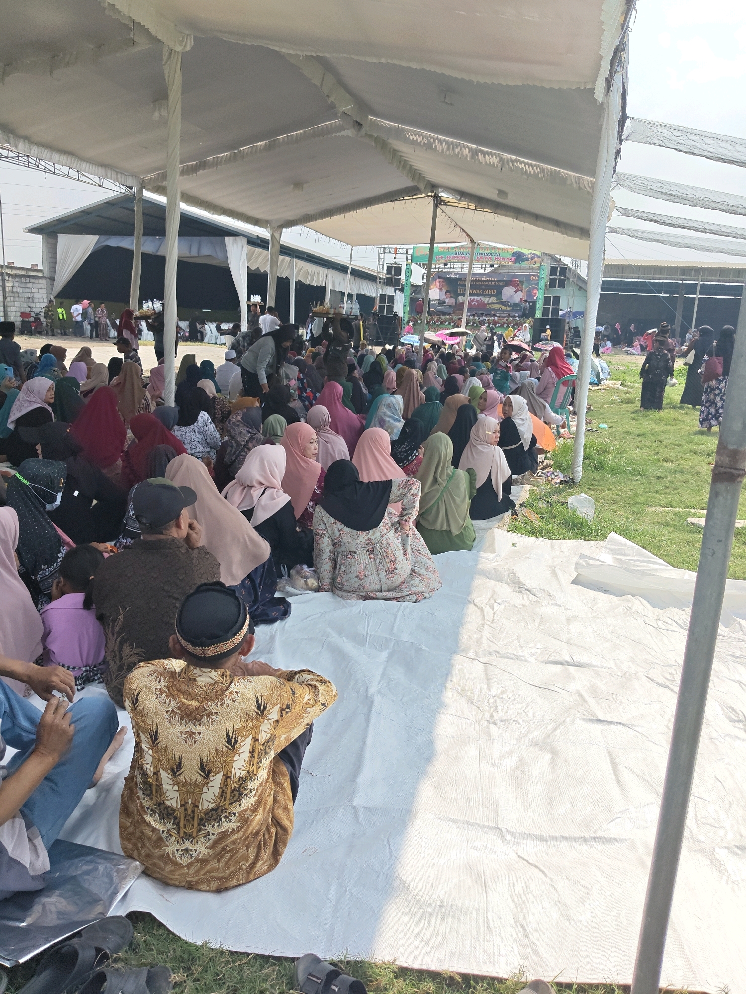 The image shows an outdoor gathering under a large white tent, likely in Bangsal, Indonesia, given the context. A crowd of people, predominantly women wearing hijabs in various colors, are seated on white tarps and the grass. In the foreground, a man wearing a patterned brown shirt and a black peci hat is seen from behind, facing away from the camera. To his left, another person's legs are visible, clad in blue jeans. The tent provides shade for the attendees. In the background, a stage with banners and speakers is set up, suggesting a formal event or religious gathering. The overall scene depicts a communal event taking place during daylight hours, with a mix of seated individuals and activity in the background.