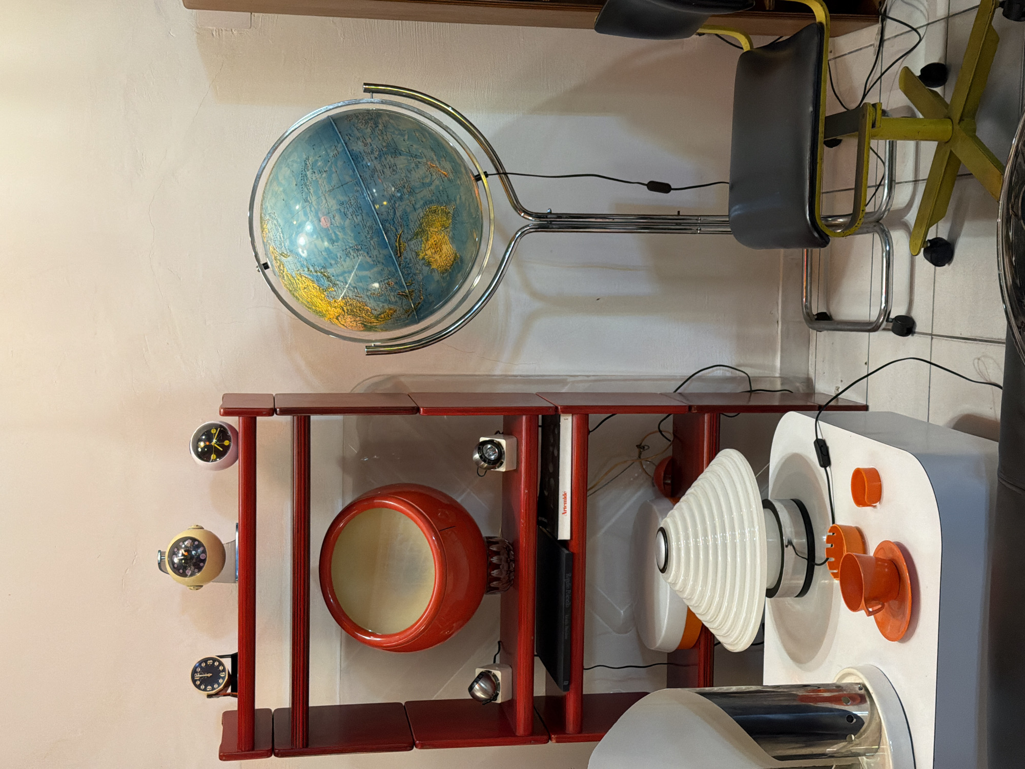 The image shows a display of vintage and mid-century modern furniture and decor items. A globe on a chrome stand is visible at the top. Below, a red shelving unit displays various items, including a red ceramic pot, clocks, books, and wall lamps. To the right, there are pieces of white and orange sculptural furniture, potentially lighting fixtures or decorative objects. The background is a plain wall. The location could be a showroom, vintage store, or an exhibition space.
