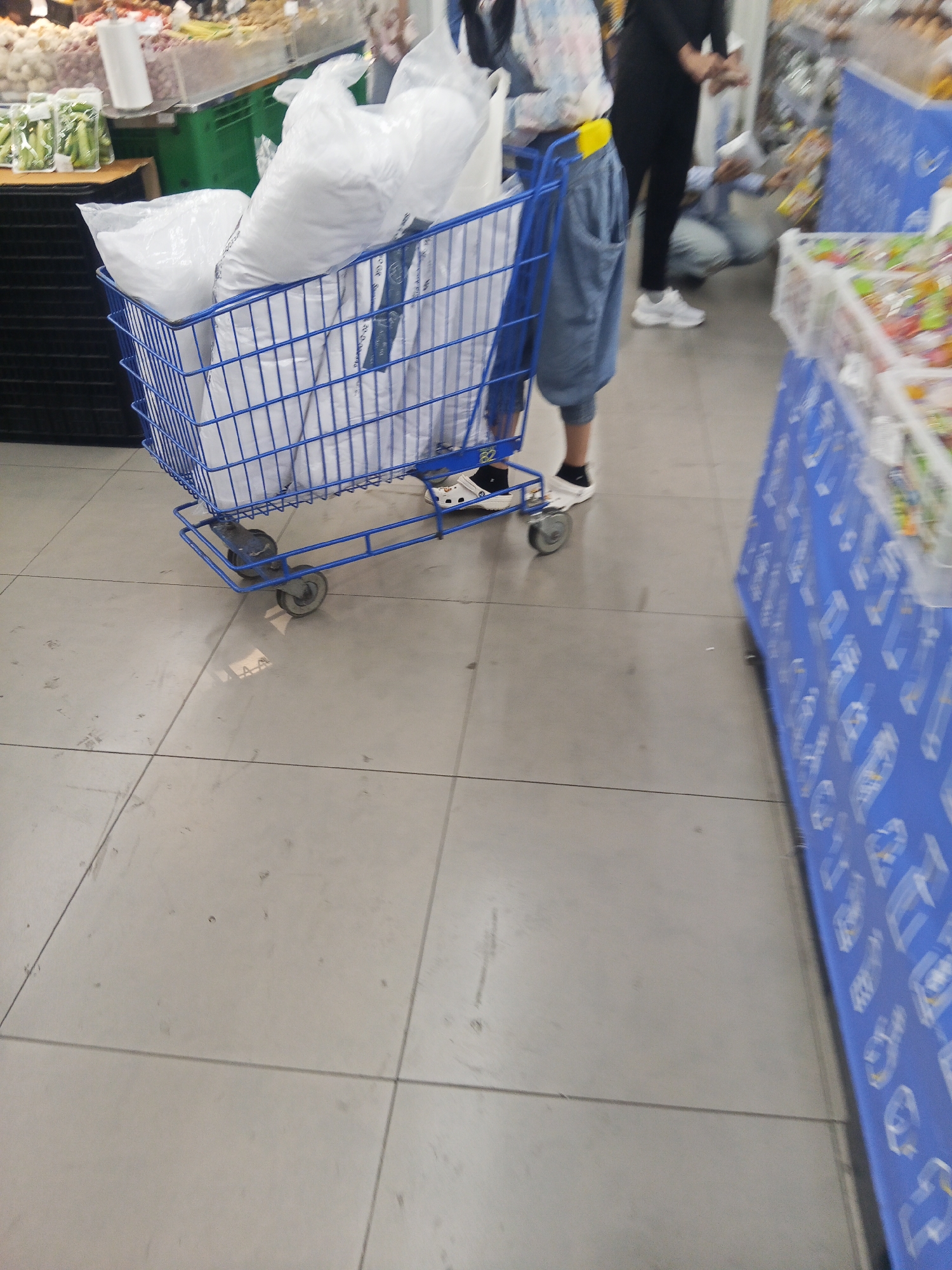 The image captures a close-up view of a blue shopping cart filled with white plastic bags. The cart has a metal frame and four wheels, two of which are visible. The bags appear to be large and opaque, suggesting they contain substantial items. The cart is positioned on a tiled floor, likely in a commercial setting. The lighting suggests an indoor environment, possibly a supermarket or a store. The floor tiles are light grey and arranged in a grid pattern. The overall scene is devoid of any clear identifying landmarks for Lubuklinggau, Indonesia, as the image focuses primarily on the shopping cart and its contents. No people are clearly depicted in focus, though blurry figures are visible in the background, suggesting a public space. The context provided by the question about Lubuklinggau, Indonesia, is not visually supported by the image itself.