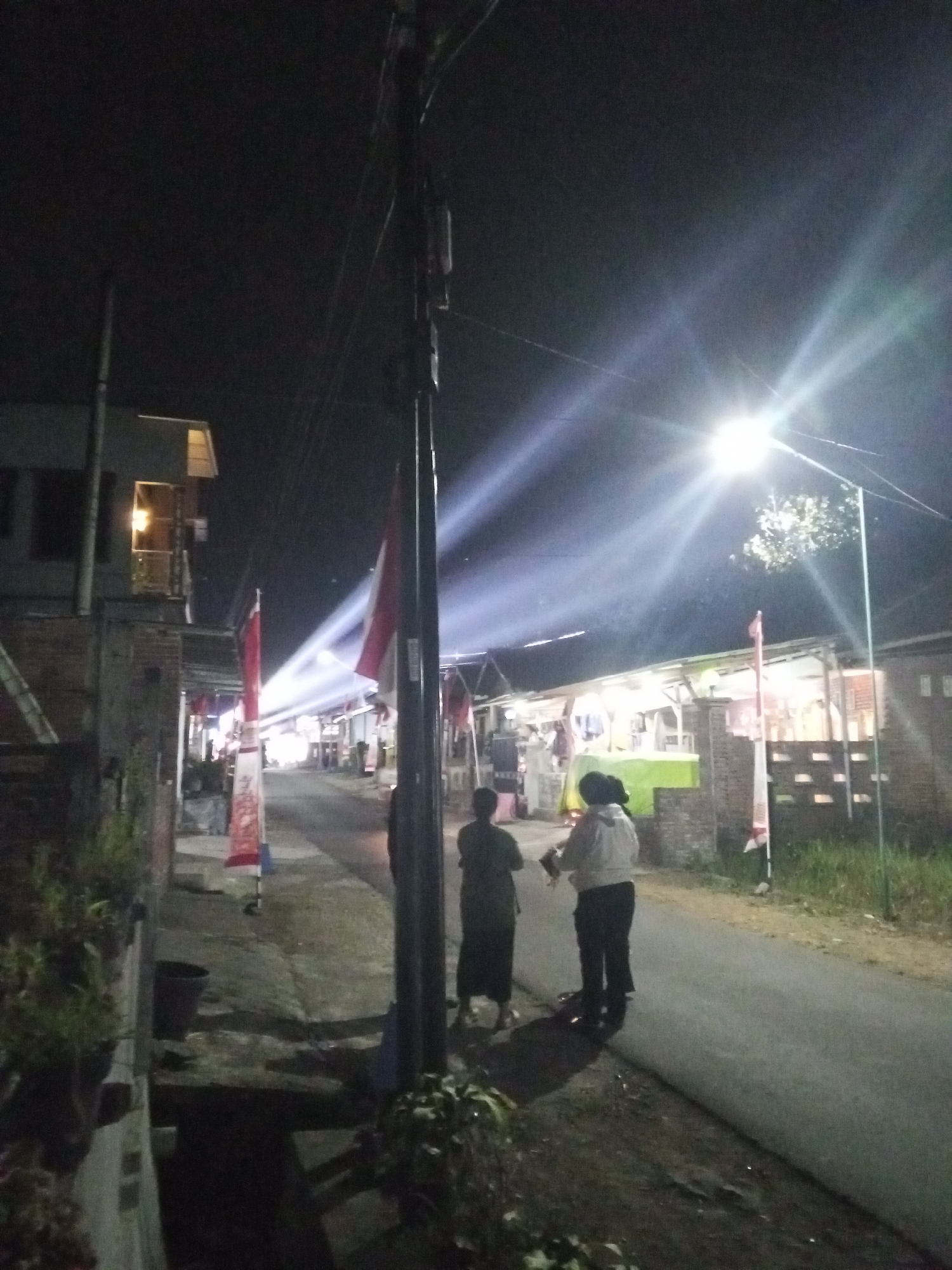 A night scene captures a street in Turen, Indonesia, likely during an event or celebration, as indicated by the prominent display of red and white Indonesian flags lining the road. A bright street lamp illuminates the scene, casting strong beams of light that create dramatic streaks across the dark sky, suggesting the presence of fog or smoke.
In the foreground, on the left side of the street, a building with exposed brick and a second-story balcony with a lit window is visible. A large black pot sits near the building's base.
On the right side of the street, several people are present. Two individuals stand near a lamppost. One person, wearing a dark garment, stands with their back to the camera. Beside them, another person wears a light-colored jacket and dark pants, appearing to hold something. Further down the street, more people are visible near stalls or small buildings, some of which are illuminated from within, with goods or decorations potentially displayed. The street itself is paved and appears to be a typical local road. The overall atmosphere is illuminated by artificial light against the dark night sky, with the flags adding a festive touch.