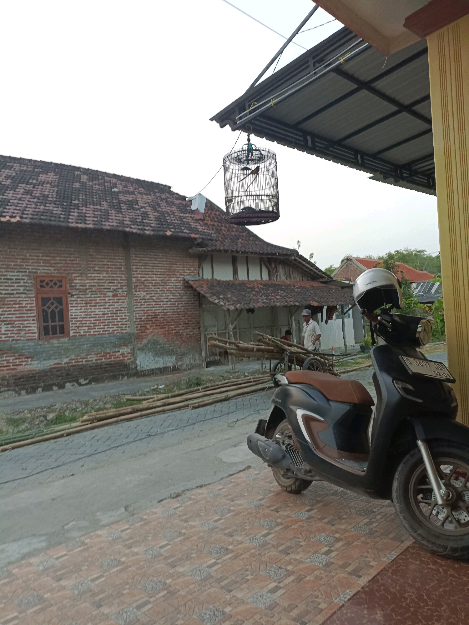 This image depicts a scene in the city of Rancang Kencono, Indonesia. A dark-colored scooter with a brown seat is parked in the foreground on a tiled pathway. A silver helmet rests on the handlebars of the scooter. To the left of the scooter, a birdcage hangs from a structure, containing at least two birds. Behind the scooter and birdcage, a traditional Indonesian house with a tiled roof and brick walls is visible. A man in a light-colored shirt and cap stands near a cart laden with bamboo poles. Another person is partially visible behind the cart. The environment appears to be a rural or semi-urban setting with trees and other buildings in the background. The sky is overcast.