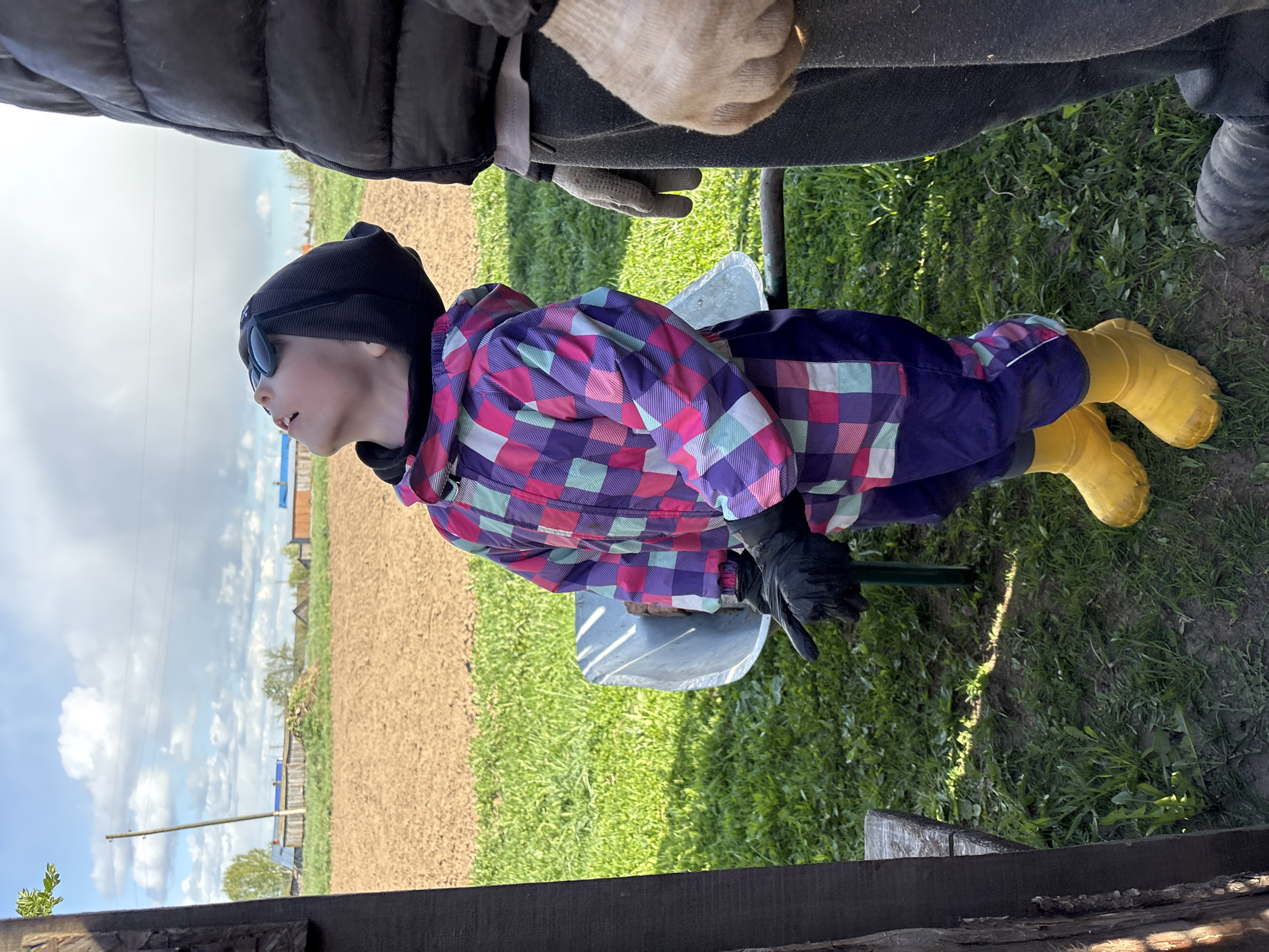 A child sits on a metal seat outdoors in Ваньки, Russia, wearing a black beanie, sunglasses, a checkered patterned snowsuit, black gloves, and yellow rubber boots. A person wearing a black jacket, grey pants and tan gloves stands above the child. The scene includes a tilled field in the background under a partly cloudy sky. The ground surrounding the child is covered in green grass and dirt. A dark wooden structure is visible in the lower portion of the frame.