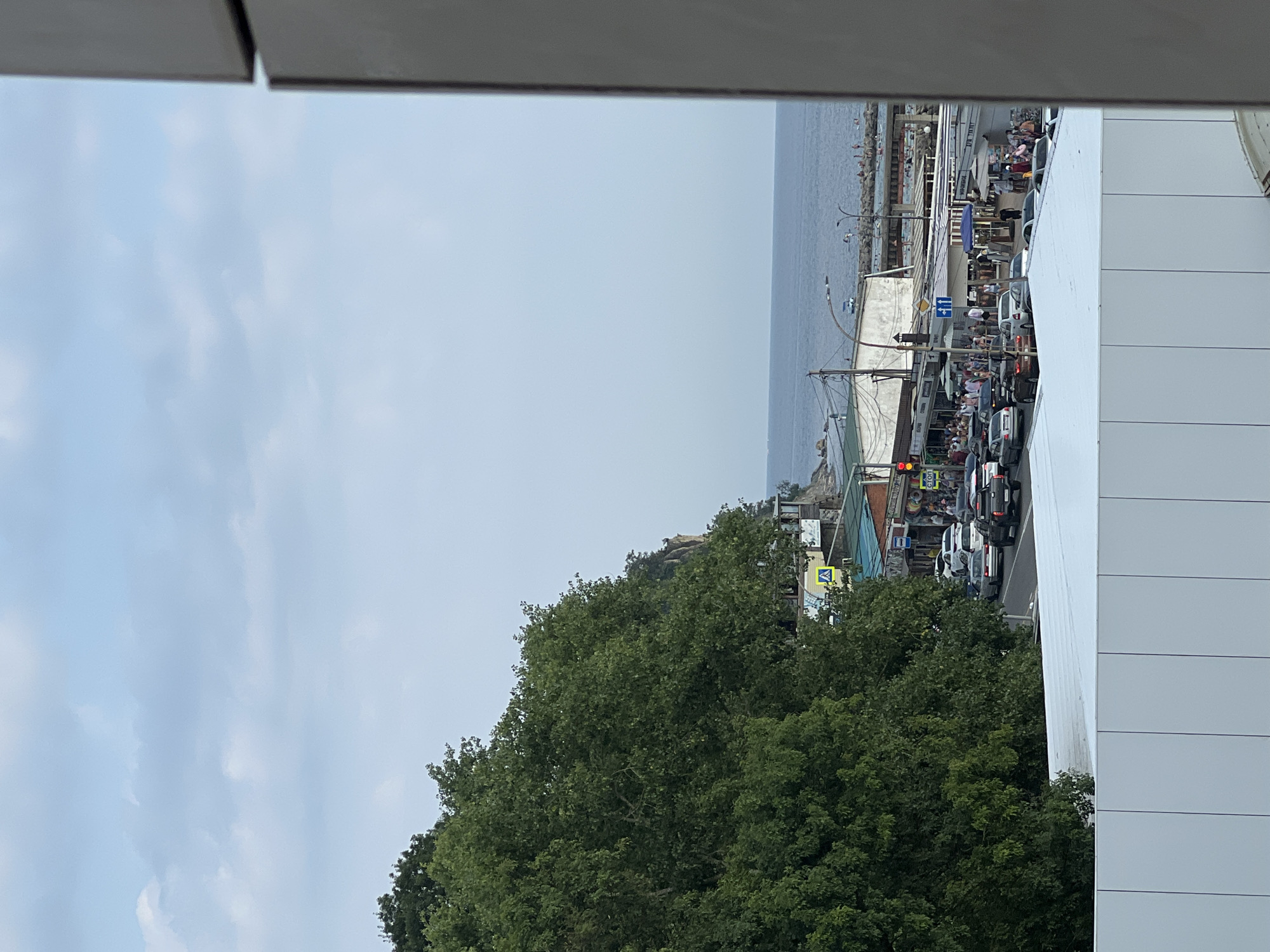 This image captures a view of the seaside city of Lermontovo, Russia, on a cloudy day. In the foreground, a white paneling structure partially obstructs the view. To the left, a dense, green tree line covers a hillside. Further into the scene, a road runs parallel to the coast, with several cars driving and parked. Along the road, a strip of buildings with awnings and shops are visible. People are seen walking along the sidewalk and on a pier extending into the calm, blue sea. The sky above is a pale blue with scattered clouds.