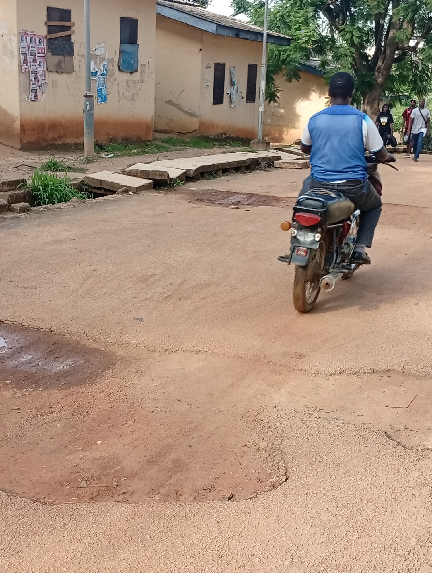 A man wearing a blue and white t-shirt and dark pants rides a motorcycle on a dirt road. The man is viewed from behind, and the motorcycle is moving away from the camera. Several other people are visible in the background, walking on the road. A building with a light-colored facade and dark windows stands to the left of the road. The environment appears to be a rural or semi-urban area, possibly within the city of Rafin Par, Nigeria, indicated by the red earth of the road and the style of the building. There is vegetation on the left side of the road, and some debris on the ground.