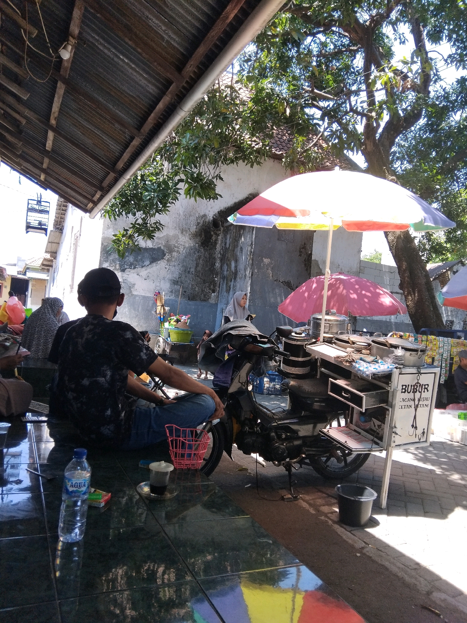 In the city of Kepuh Kiriman, Indonesia, an outdoor scene unfolds under a corrugated metal roof. A person wearing a black t-shirt and jeans sits with their back to the camera, facing a mobile food stall. The stall, attached to a motorcycle, is adorned with a colorful umbrella and a pink umbrella. It displays pots, bowls, and a sign that reads "BUBUR KACANG HIJAU KETAN HITAM" (Mung Bean Porridge Black Glutinous Rice). A bottle of Aqua water and a pack of cigarettes are on the tiled floor next to the person. In the background, other individuals are visible, some seated and others standing. A tree with lush green leaves shades the area.