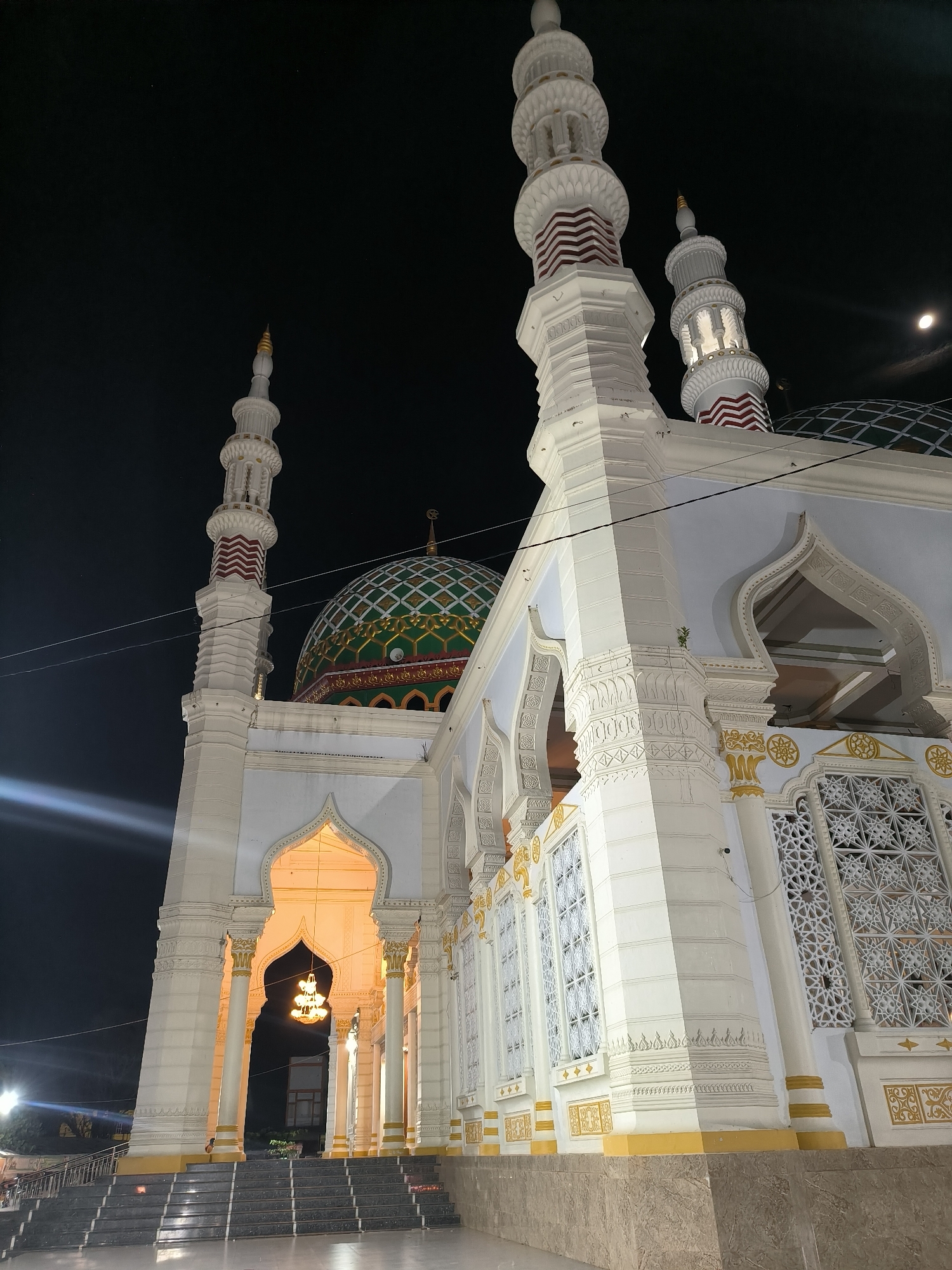 At night, the Baitul Makmur Grand Mosque stands prominently in Kotapantonlabu, Indonesia. The mosque's exterior is predominantly white with gold and red accents, featuring intricate geometric designs and patterns. A large green and gold dome tops the structure, flanked by tall minarets. The entrance is illuminated, revealing a chandelier hanging within. Steps lead up to the entrance, and a faint light source is visible in the background. The night sky is dark, with a visible moon.