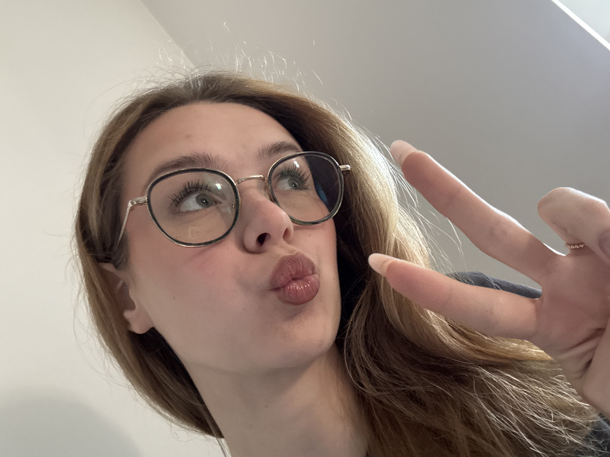 A young woman is posing for a selfie, holding up a peace sign. She has long brown hair and is wearing round glasses. Her lips are puckered. The background is a plain white wall. The location is unknown and there is nothing in the image that indicates it is Kragujevac, Serbia.