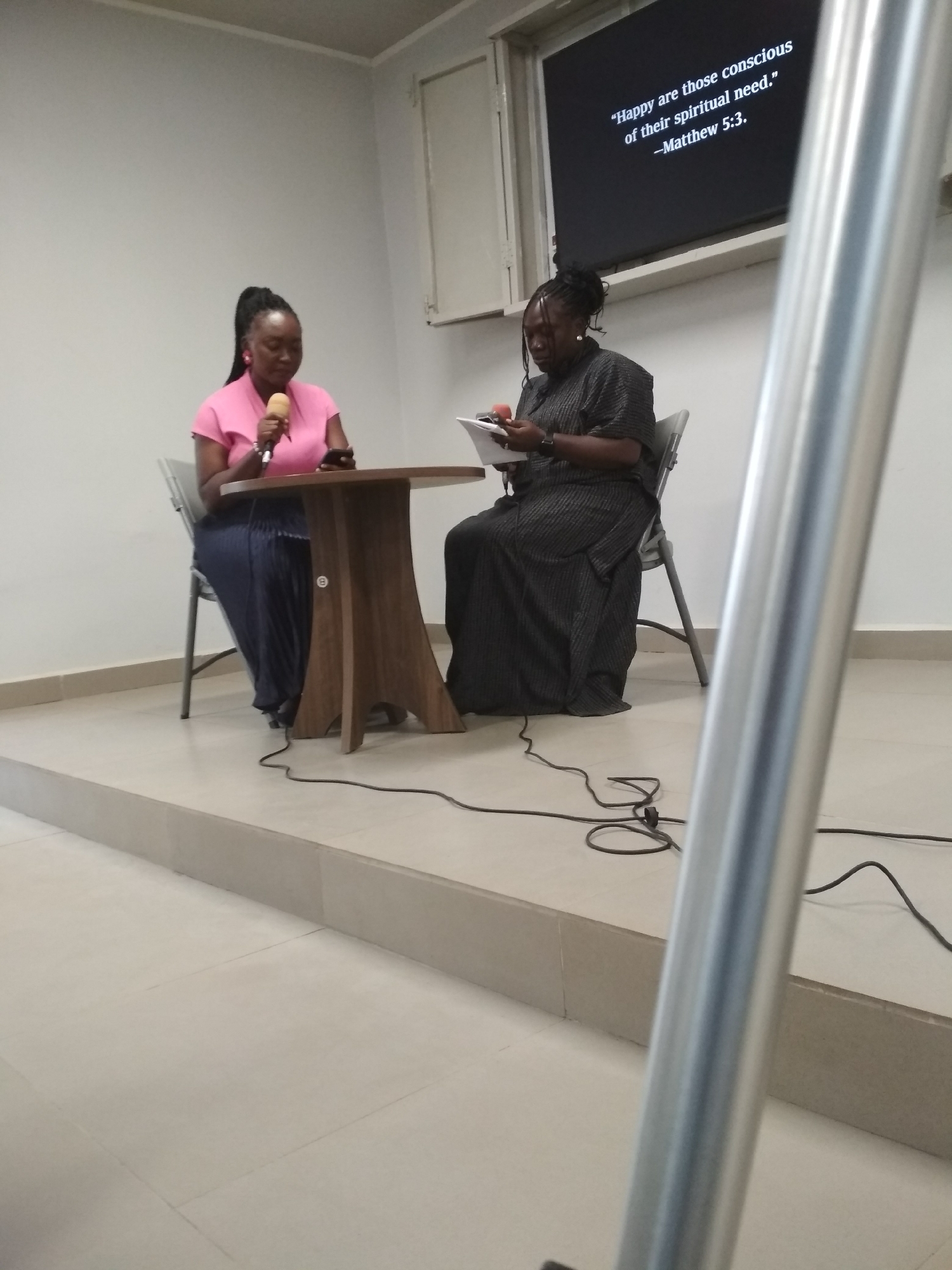 Two African women are sitting at a small, round table on a slightly elevated platform in a room. The woman on the left is wearing a pink top and a long, dark skirt. She is holding a microphone in her right hand and looking down at what appears to be a phone in her left hand. The woman on the right is wearing a dark, striped dress and looking at a document in her hands while holding a microphone. Behind them is a wall, and above the woman on the right is a screen displaying the text: "Happy are those conscious of their spiritual need. -Matthew 5:3." Cables from the microphones are visible on the floor. The room appears to be simply decorated. The exact location in Gura Top, Nigeria, is impossible to determine from the image.