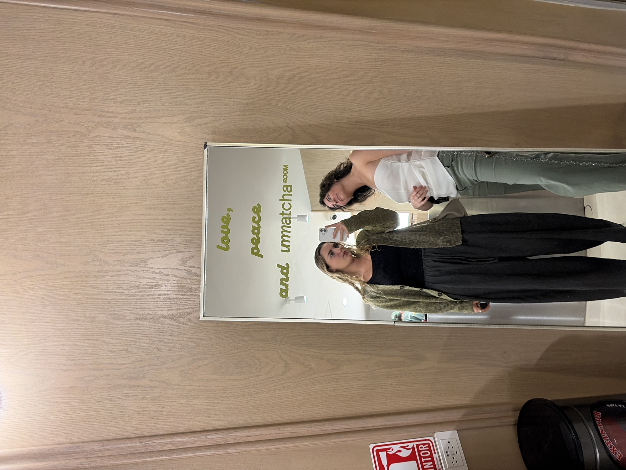 The image is a reflection in a mirror showing two women in what appears to be a changing room. The mirror has text on the glass reading "love, peace and unmatcha room." One woman is wearing a white top and green pants, while the other is in a black top, dark pants, and a green cardigan, holding a cell phone to take a picture. The background shows a wall with wooden texture. Near the bottom of the frame, a fire extinguisher sign and a can are visible. It's not possible to determine the exact location in Puebla, Mexico based solely on this image.