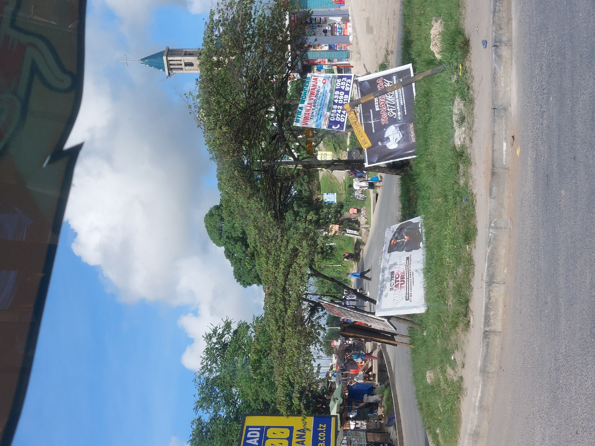 The image shows a street scene in Dar es Salaam, Tanzania. There are advertisements posted along the side of the road, advertising land for sale and a Saturday performance. The ads display phone numbers and prices in Tanzanian Shillings (TSH). In the background, a church tower is visible. The street has a mix of people and street vendors. The weather appears sunny with some clouds. The image is taken from a vehicle, with a portion of the vehicle's interior visible in the frame.