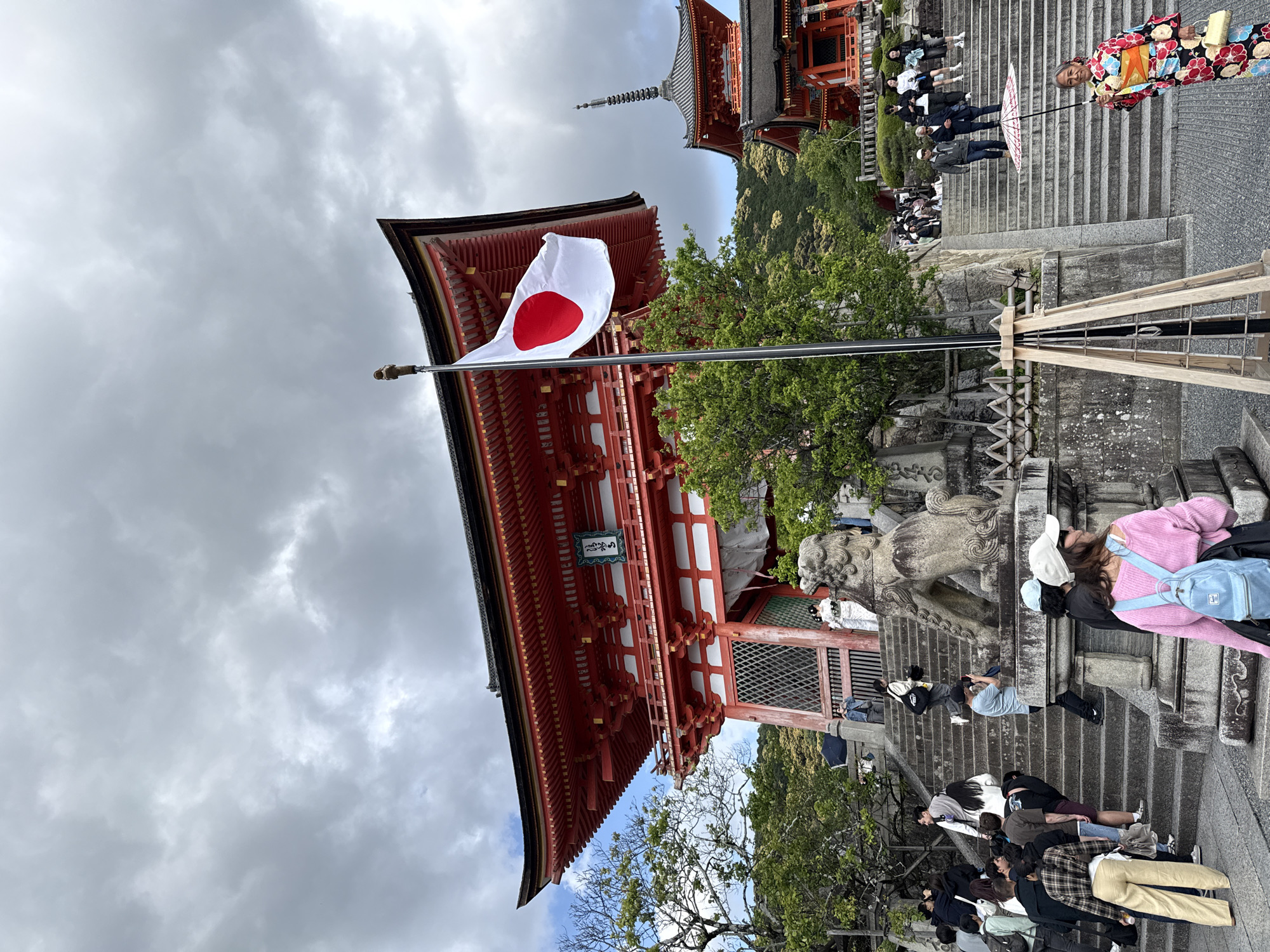 The image shows a view of Kiyomizu-dera Temple in Kyoto, Japan. A Japanese flag is prominent in the foreground, positioned in front of the red-lacquered main hall. The sky is overcast with grey clouds. Groups of tourists are seen walking up and down the stone stairs leading to the temple. A woman in traditional Japanese clothing is seen with an umbrella on the upper part of the stairs. A pagoda is visible in the background. The surrounding area features trees and stone sculptures.