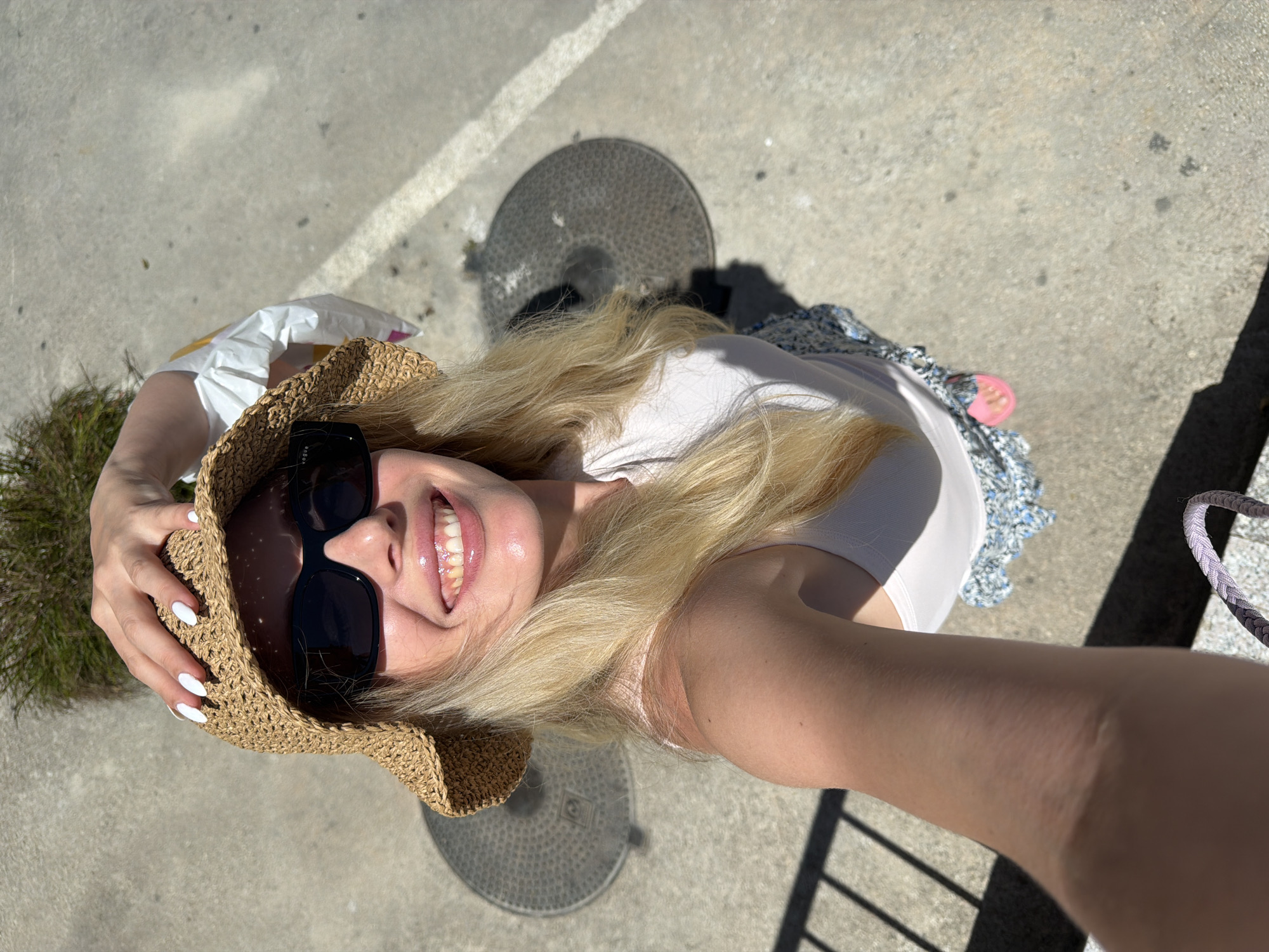 A sunlit, high-angle shot depicts a fair-skinned woman outdoors. She is wearing a straw hat, large dark sunglasses, and a light-colored top. Her long blonde hair is loose around her shoulders. Her manicured nails are painted white. She is smiling broadly, revealing her teeth.
The woman is standing on a concrete surface with two circular manhole covers nearby. A small patch of greenery is visible in the upper left corner. A portion of a railing is visible on the lower right.
Without further information, it is not possible to definitively place the location as Budva, Montenegro.