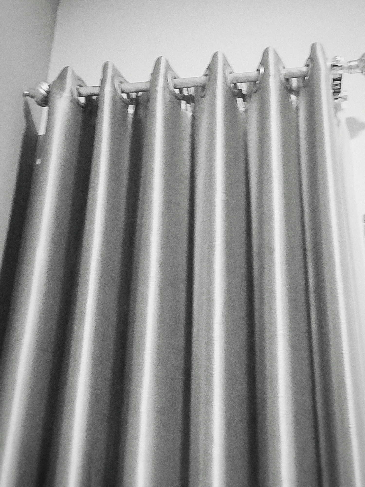 The image shows a close-up, black and white view of a set of fabric curtains hanging on a rod. The curtains are pleated, with vertical folds creating a textured appearance. Light reflects off the fabric, highlighting the pleats and the sheen of the material. The curtain rod is visible at the top, and the curtains appear to be made of a smooth, possibly satin-like material. The background is a plain, light-colored wall. No people, actions, or interactions are depicted. The scene is static and focuses on the details of the curtain's texture and hanging mechanism. The location in Mojoagung, Indonesia is not discernible from the image itself.