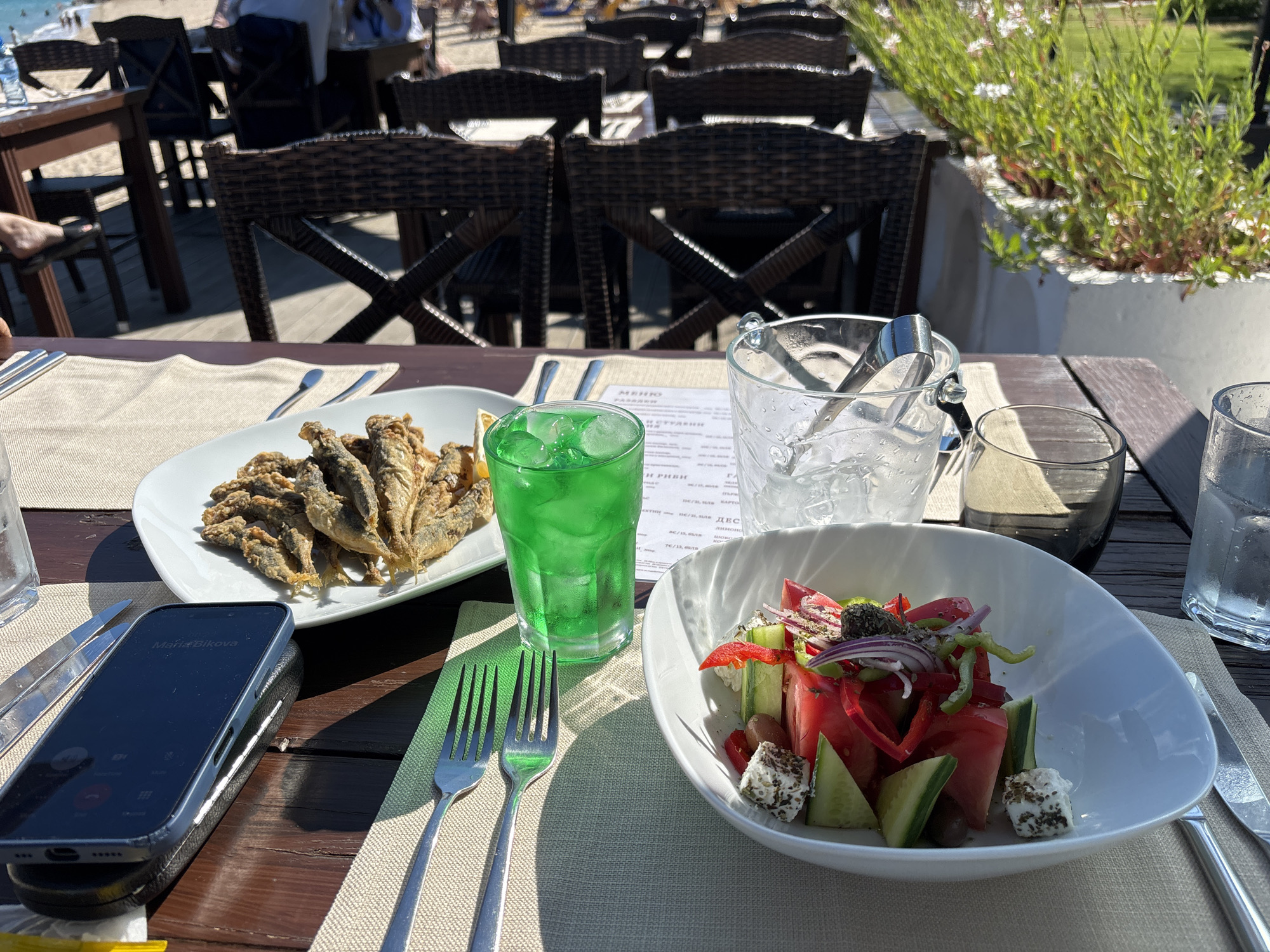 This image depicts a table set for a meal at an outdoor restaurant. The table is made of dark wood planks and covered with a beige placemat. In the foreground, there is a white bowl filled with a Greek salad, containing tomatoes, cucumbers, red onions, green peppers, olives, and feta cheese. To the left of the salad, there is a white plate with a serving of small fried fish. A tall glass with a green beverage and ice is placed next to the fish. An ice bucket filled with ice and tongs is also visible. On the left side of the table, a smartphone is lying face up on its case. Utensils, including forks and knives, are arranged on the placemat. In the background, several wicker chairs are visible around other tables, and beyond that, a beach with people and the sea can be seen. The overall setting suggests a casual dining experience near the coast. The image was taken in Varna, Bulgaria, likely at a restaurant located along the beachfront.
