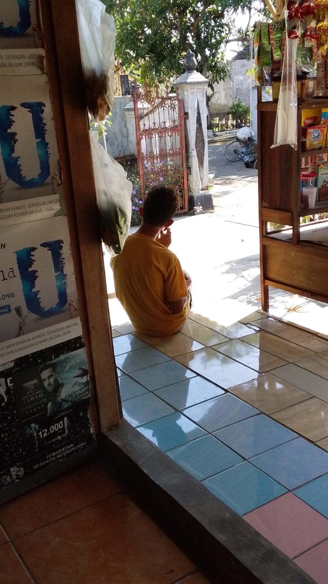 A young male, appearing to be of Indonesian ethnicity with dark, curly hair, is seated on a tiled floor in what appears to be an open-air stall or shop entrance. He is wearing a bright yellow t-shirt and dark shorts. The man is facing away from the camera and appears to be touching his ear or face. The floor is tiled with square tiles in shades of blue and brown. To the left of the frame, there is a wooden structure with stacked posters or magazines. One poster clearly shows the letter "U" in a blue, stylized font. To the right, a wooden shelf unit displays various packaged goods, possibly snacks or toiletries, with some plastic bags hanging. In the background, there is a decorative metal gate, a white pillar, lush green trees, and a bicycle parked. Sunlight casts strong shadows on the ground. The overall scene suggests a casual, possibly commercial, outdoor setting in Lawu, Indonesia.