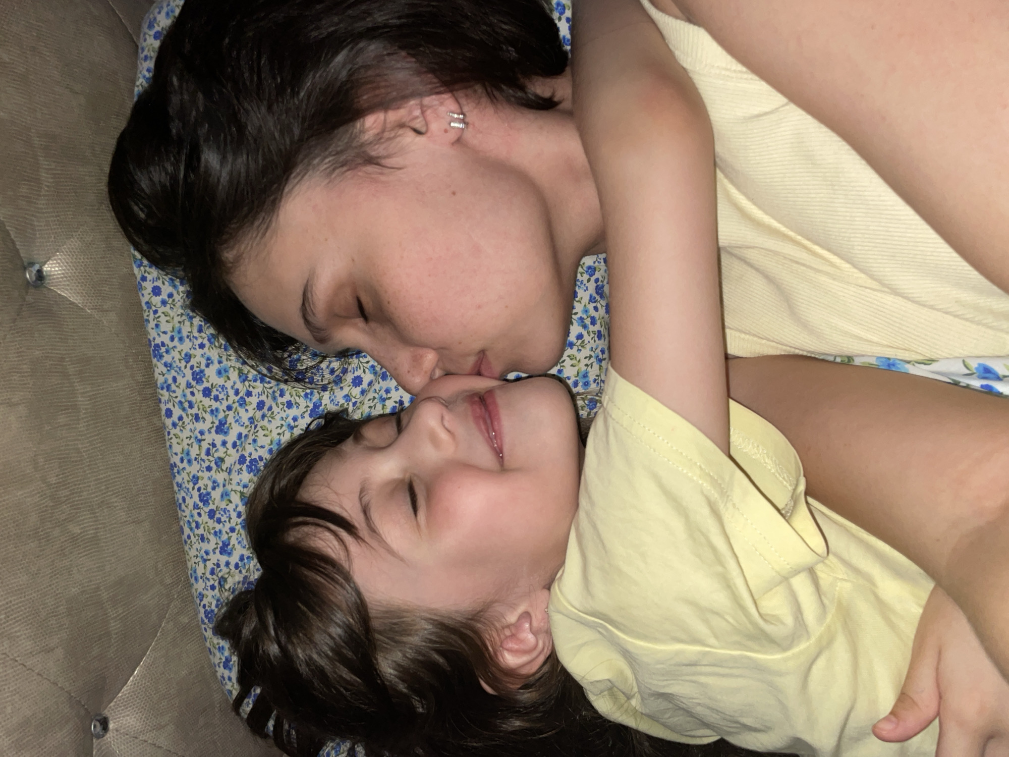 A woman and a child lie on a floral patterned pillow on top of what appears to be a textured, possibly upholstered headboard or couch. The woman has dark hair, is wearing a silver earring, and has her eyes closed. The child also has dark hair and is smiling with their eyes closed. The woman's face is close to the child's, potentially kissing them. Both are wearing light yellow shirts. There is no indication of Izhevsk, Russia or any specific location.
