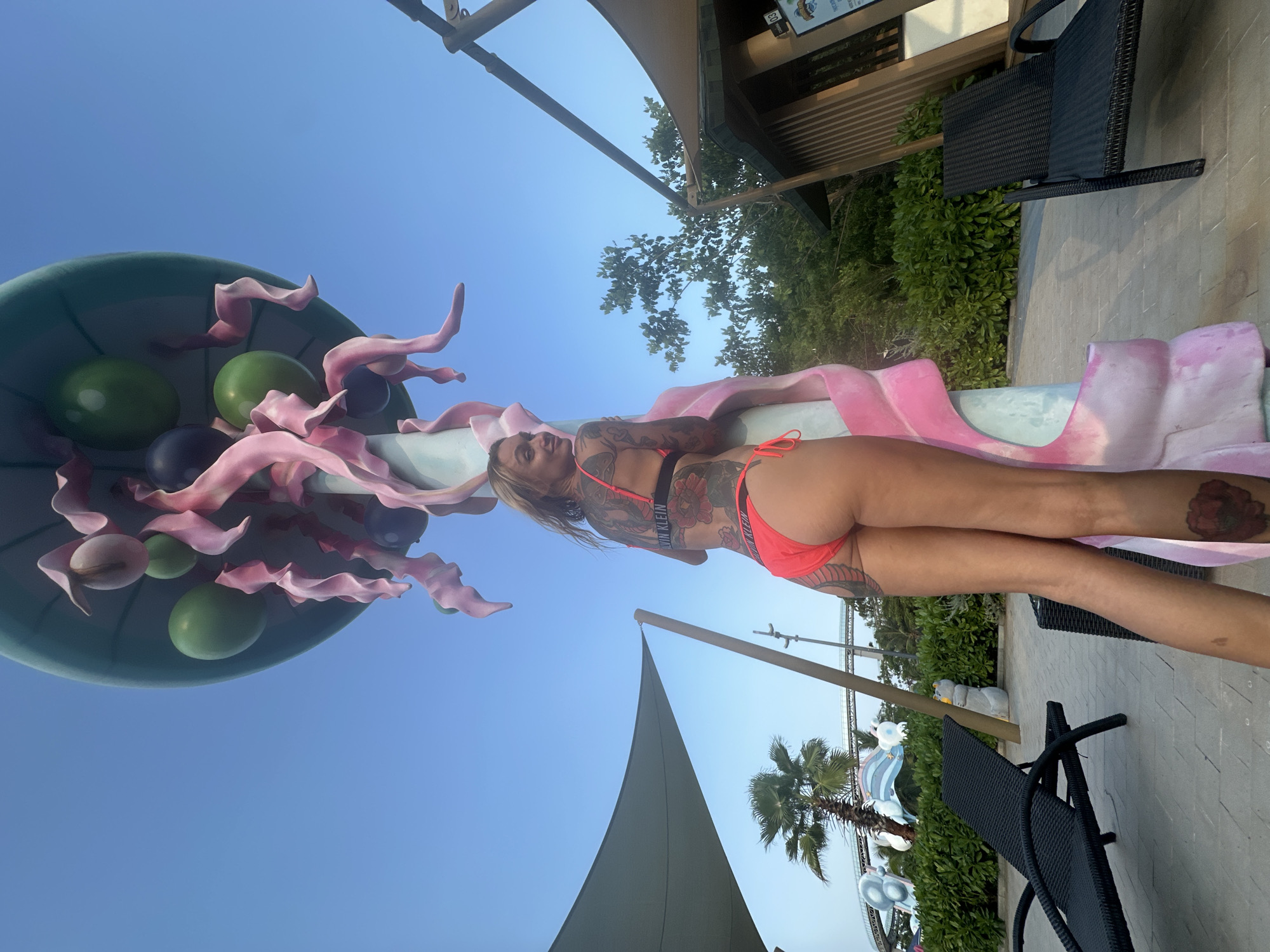 The image depicts an outdoor scene in Tien Thanh Ward, Vietnam, characterized by bright sunlight under a clear blue sky. A woman is positioned in the foreground, facing partially away from the camera, looking over her left shoulder. She has blonde hair and is wearing a coral-orange two-piece swimsuit, with "CALVIN KLEIN" visible on the band of her bikini top. Her back, arms, and legs are extensively covered with tattoos, including floral designs on her back and a large red flower on her lower right leg. She is leaning against a large, light blue pole adorned with undulating pink, organic-shaped sculptures that appear to twine around it. The pole is topped with an inverted teal cap, from which several smooth, ovoid or spherical objects in green, light pink, and dark purple/blue hues are suspended.
To the left of the woman, a large grey shade canopy is visible, supported by a pole, beneath which a black woven lounge chair rests on light grey paving stones. Further left and in the distance, a palm tree stands, and various structures indicative of a water park or resort are present, including a roller coaster-like track and stylized white and blue abstract elements. To the right of the woman, a wooden-slatted building with a sign (partially visible as "JUNGLE SPLASH") is situated behind green foliage, and another black woven lounge chair is present. The ground is paved with light grey interlocking pavers, transitioning into landscaped areas with green bushes and trees.
