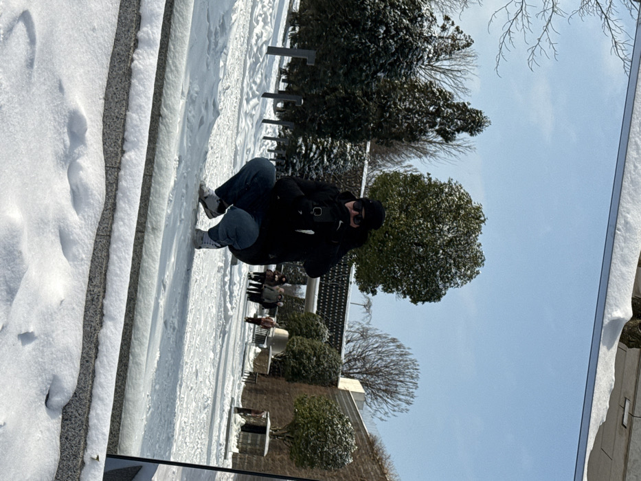 This image captures a reflection in a large, vertical mirror-like surface, situated outdoors in a snow-covered environment within the city of Plodorodny, Russia. The scene is illuminated by bright daylight under a clear blue sky.
In the central foreground of the reflection, a person is depicted in a low squatting stance. This individual wears a black winter jacket, dark blue jeans, white sneakers, a black beanie, and black sunglasses, with gloved hands. They are holding a black smartphone directly in front of their face, positioned to capture a self-portrait of their reflection.
The background, visible behind the primary subject within the reflection, features a park-like setting covered in a layer of fresh snow. To the left, several snow-laden evergreen trees and other bare-branched trees are present, alongside a row of dark, vertical posts protruding from the snow. To the right, a large, dark-textured architectural structure, possibly a wall with a central archway, spans horizontally. Further to the right, a multi-tiered stone wall with some small, rounded green shrubs is visible. Several indistinct figures, appearing to be other pedestrians dressed in winter clothing, are walking along a distant snowy path near the architectural structure.
The reflective surface is bordered by a narrow metallic strip at the top and rests upon a low, snow-covered stone ledge at the bottom. The photograph capturing this reflection is taken from a low vantage point, looking upwards at the reflective surface.