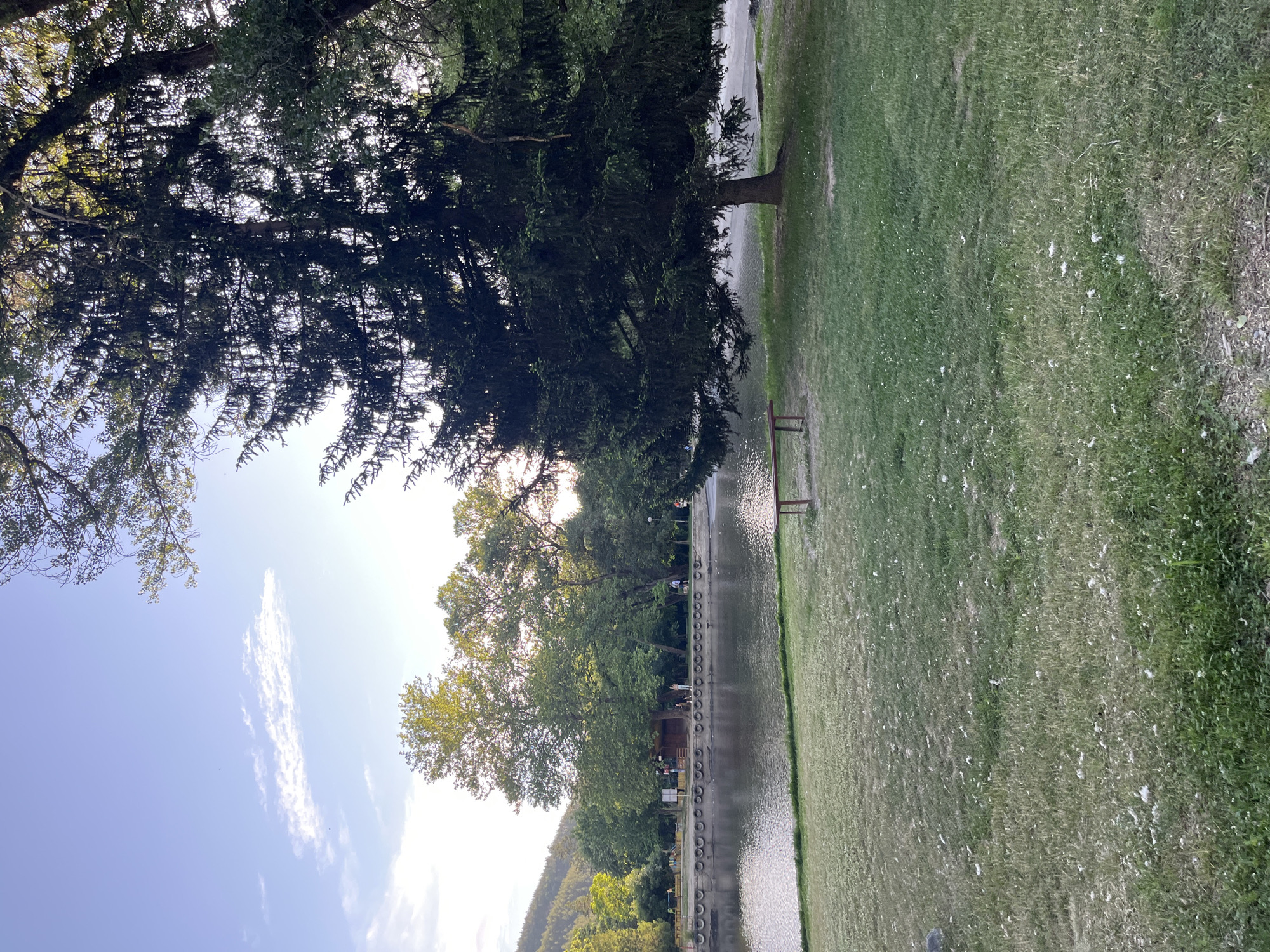 The image shows a park in Blagoevgrad, Bulgaria. The scene includes a body of water, possibly a lake or large pond, with a tire-lined edge. Green grass covers the ground, and trees are prominent. There's a bench visible near the water's edge. In the background, a low mountain can be seen. The sky is blue with scattered clouds. The lighting suggests it's daytime.