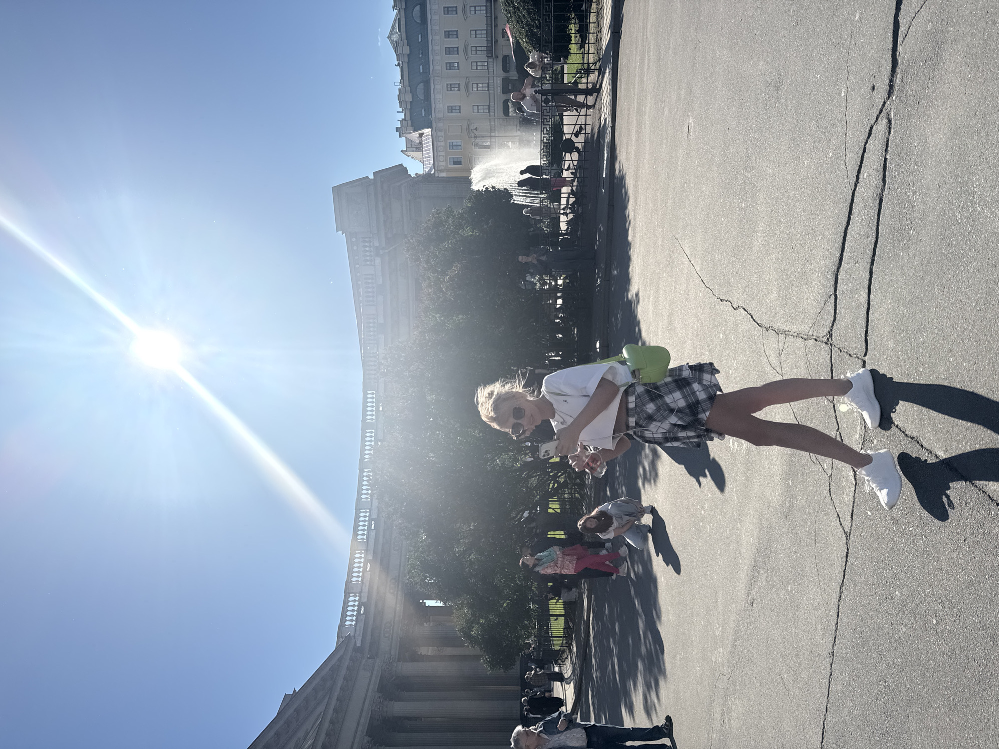 This media file depicts an outdoor scene in Saint Petersburg, Russia, on a bright, sunny day. The sun is a prominent feature, casting strong lens flares and sharp shadows across the pavement, indicating it is likely midday. The image shows a paved area, possibly a plaza or walkway, with distinct cracks in the asphalt.
The primary subject in the foreground is a young woman wearing sunglasses, a white t-shirt, and a plaid skirt. She is holding a smartphone in one hand and what appears to be a drink in the other. Her stance suggests she is either taking a photo or interacting with her phone. Beside her, and slightly out of focus, is a light green object, possibly a bag or accessory.
In the midground, several other individuals are visible. Some are walking, while others appear to be gathered near a fountain. The fountain is actively spraying water, suggesting it is operational. Behind the fountain is a lush green area with trees, and further back, several multi-story buildings with classical architectural elements can be seen. These buildings, with their distinctive windows and facades, are characteristic of Saint Petersburg.
The overall atmosphere appears to be relaxed and leisurely, with people enjoying the sunny weather outdoors. The presence of the fountain, the architecture, and the general ambiance are strong cues for the location being Saint Petersburg. No visible text is present in the media file.