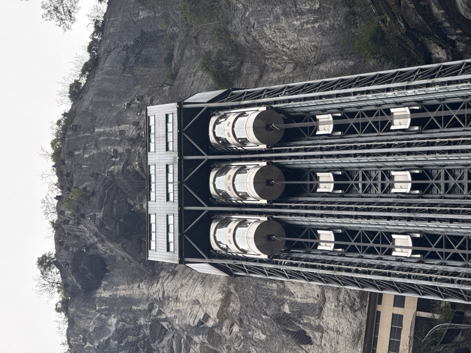 The image depicts a colossal outdoor panoramic elevator structure situated against a towering, steep grey rock cliff face in Wulong District, China. The primary structure features a dark grey metallic truss framework, housing three parallel vertical elevator shafts. Each shaft contains a cylindrical, glass-enclosed elevator car, visible at varying heights. Below these main cabs, smaller, white rectangular components are visible further down each shaft. The upper section of the elevator complex consists of a multi-story building component with multiple horizontal windows.
The adjacent cliff face is extensively covered with a grey metal mesh netting, secured by numerous square anchors, indicating rockfall protection measures. Sparse green vegetation and bare trees grow on the cliff edges and top. To the lower left, a section of a lighter-colored building with rectangular windows is partially visible at the cliff base. The sky above is uniformly overcast and light grey. No people, specific actions, or interactions are discernible within the frame.