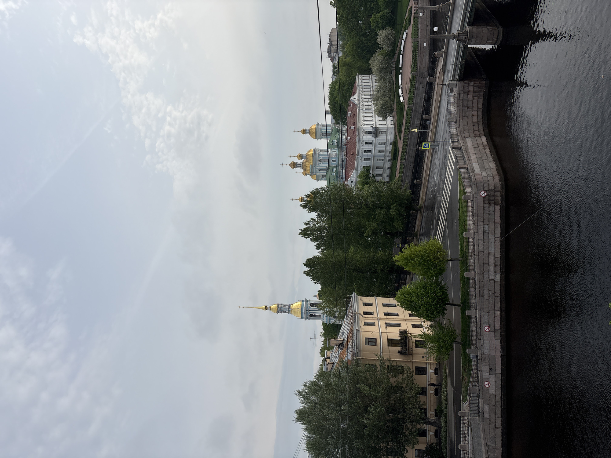 This image depicts a view in Saint Petersburg, Russia. It shows a canal or river with dark water, bordered by a stone embankment. A road runs along the embankment, marked with a zebra crossing. Buildings, including a church with golden domes and a tall spire, are visible among green trees. The sky is overcast with scattered clouds. There are no discernible people or actions taking place in the frame.
