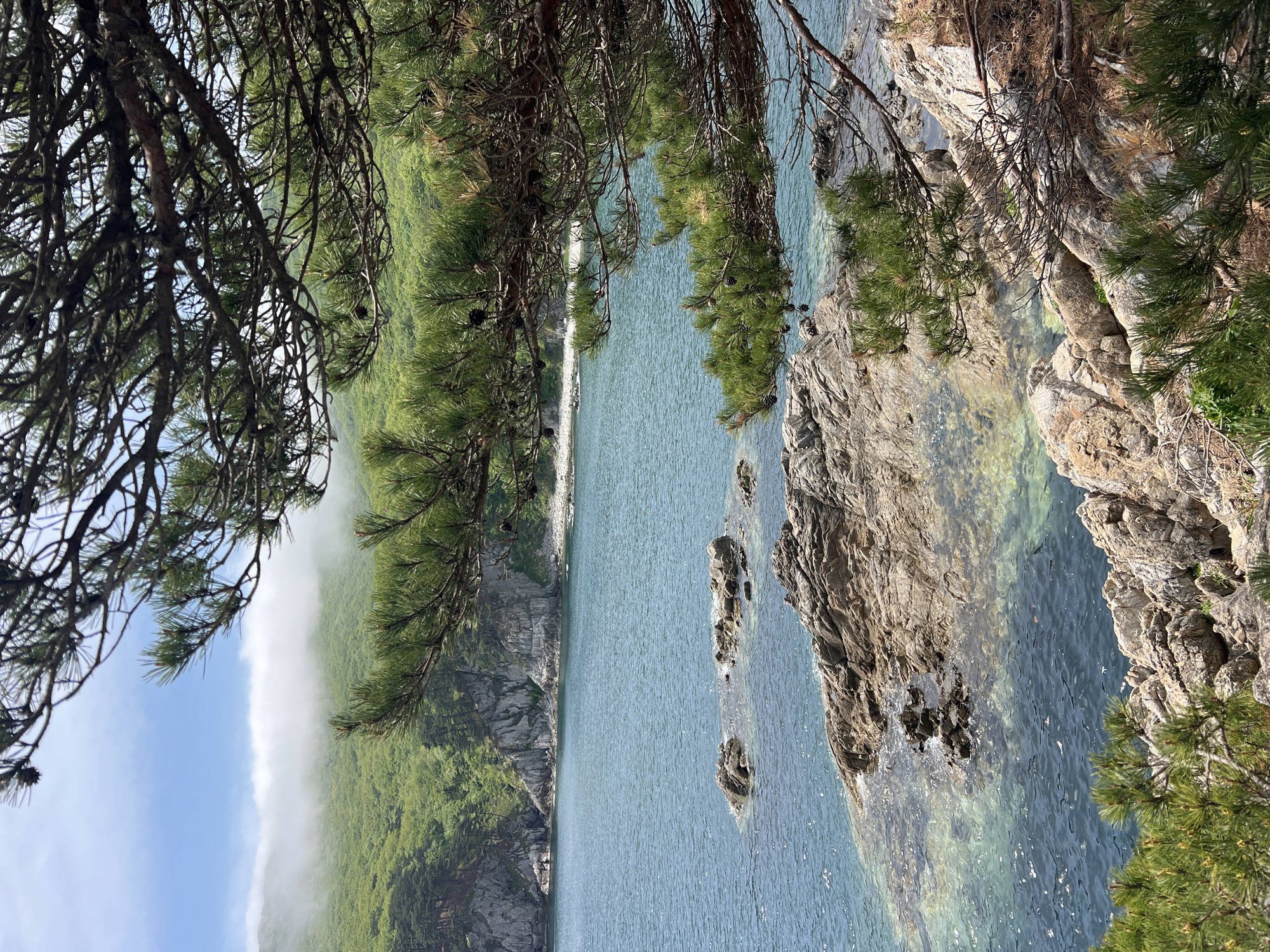 The image shows a view of Mayak Gamov, Russia. The scene is dominated by the landscape, featuring a forested cliffside meeting a body of water. The water is light blue and has several rocks that protrude from it. The vegetation consists primarily of green trees. The composition is framed by tree branches in the foreground. On the far left, a white fog extends from the top of a mountain to the sky.