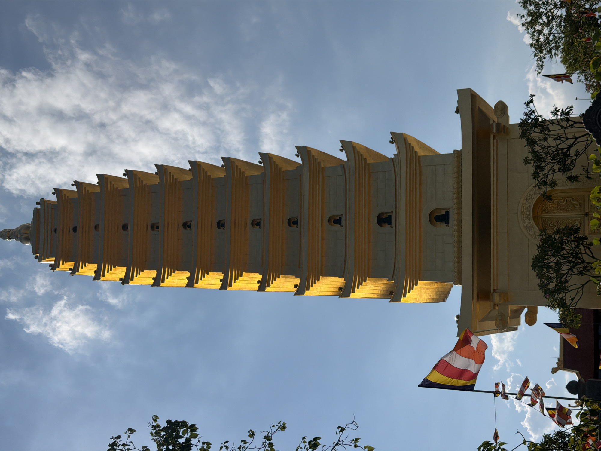 A low-angle shot shows the towering stupa of Long Son Pagoda in Nam Nha Trang Ward, Vietnam, against a blue sky with scattered clouds. The stupa is multi-tiered, with each tier featuring a projecting roof and arched openings. The structure is painted in light yellow and tan, with the setting sun casting highlights and shadows on its surface. A string of Buddhist flags in various colors hangs near the base of the stupa, visible among trees.