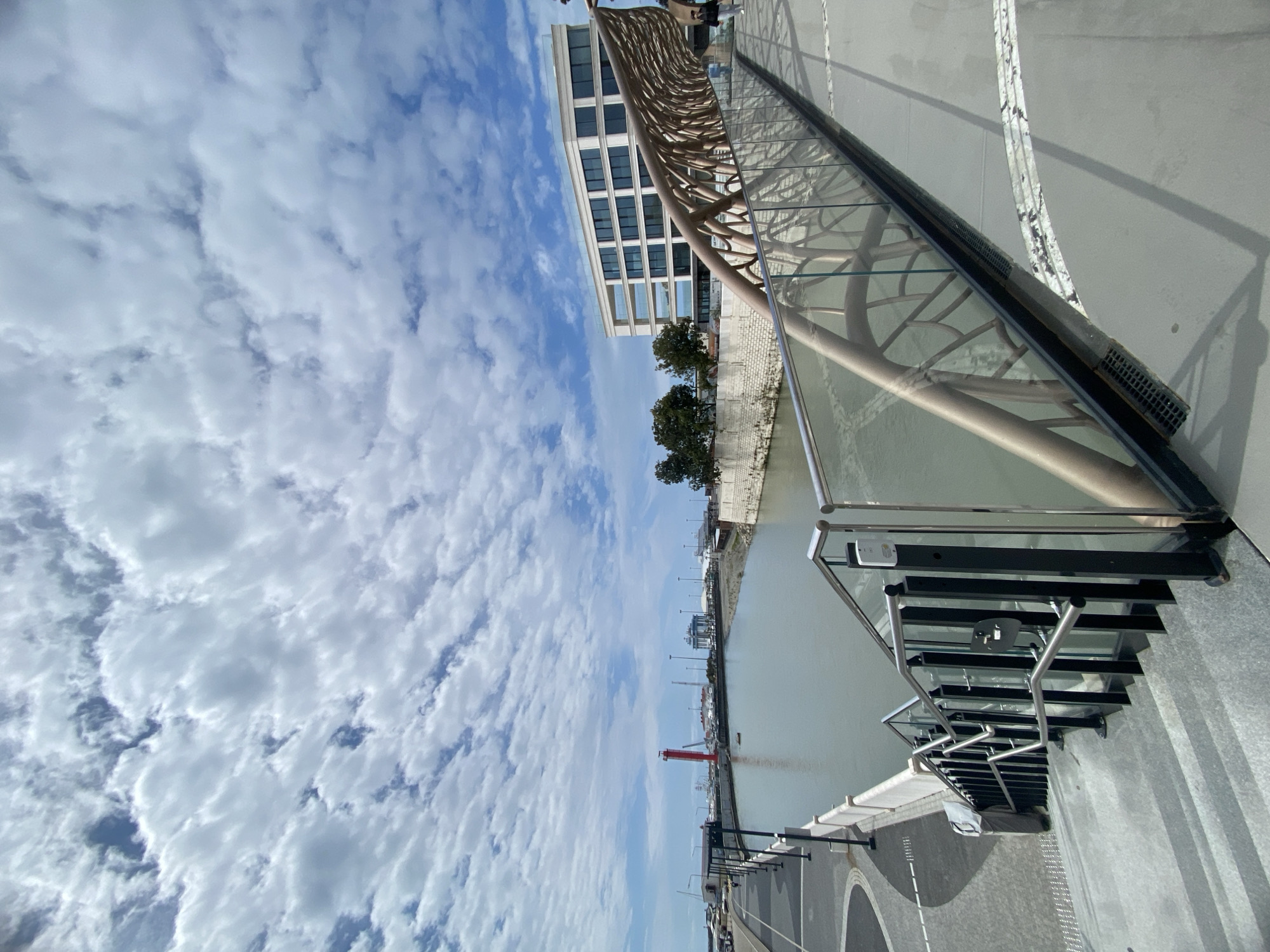 This image captures a bright, modern waterfront scene in Sochi, Russia, likely a canal or river alongside a promenade. The sky dominates the upper portion, featuring a clear blue background dotted with numerous scattered white and light grey cumulus clouds, suggesting fair weather.
Running parallel to the water is a distinctive architectural structure with an organic, lattice-like design in a light tan or brown color, resembling intertwined branches. This structure appears to be part of a pedestrian bridge or an ornate railing system. Behind this, on the far bank of the water, stands a contemporary multi-story building with a light-colored facade and numerous windows. A few green trees are visible along the water's edge, and in the distance across the water, various urban elements including streetlights and a prominent red crane or industrial structure can be seen. The water itself is calm and reflects a muted greyish-green hue.
In the foreground and to the right, a wide, paved promenade or walkway is visible, characterized by dark and light grey sections and some white markings, possibly for pedestrian paths or parking. Modern glass and stainless steel railings line the edge of this promenade, providing a clear view of the water and the structures beyond. There are also black metal posts or fixtures along the walkway.
The scene appears peaceful and well-maintained, indicating a pleasant, sunny mid-day. There are no specific activities taking place, though a faint figure of a person is visible walking in the distance on the promenade. No discernible text is visible within the image. The overall impression is one of modern urban design integrated with natural elements, under an expansive, cloudy sky.